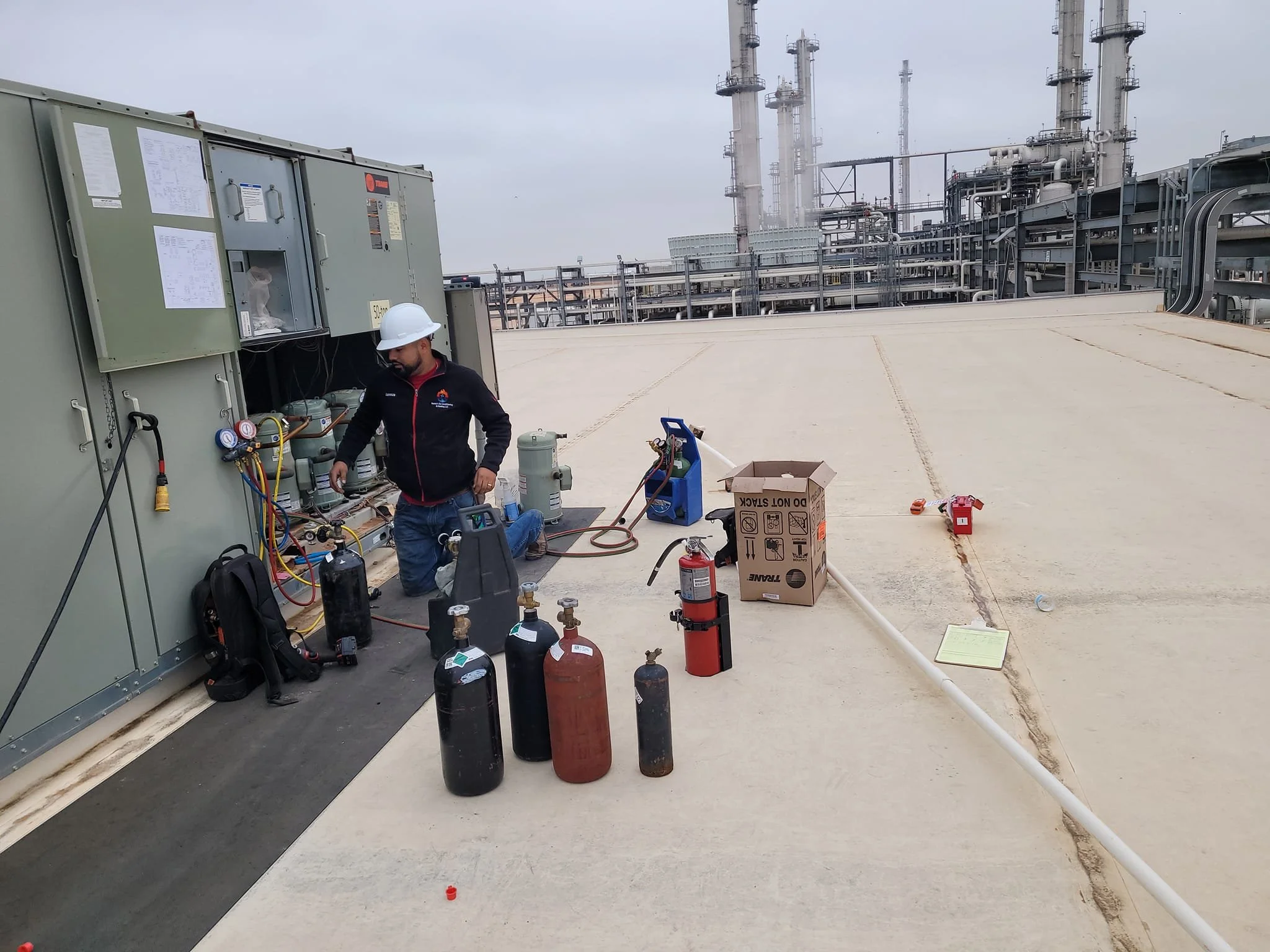 A technician wearing a white hard hat and black jacket kneels on a rooftop working with refrigeration or air conditioning equipment, with various tools, cylinders, and a cardboard box nearby, and industrial pipes in the background.