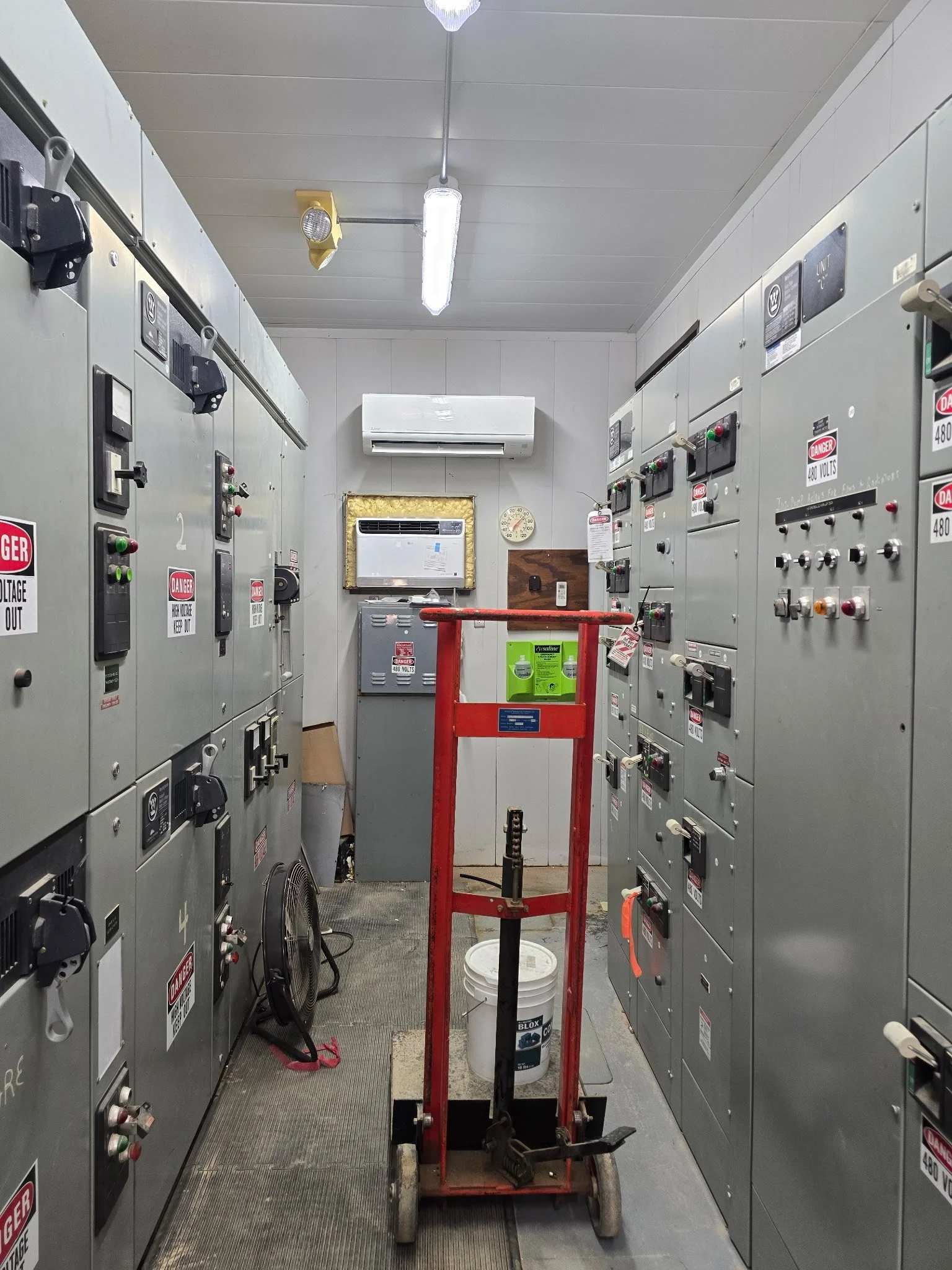 A room with electrical control panels on both sides, a red hand truck or dolly in the middle, a bucket, a fan, an air conditioner, a wall clock, and fluorescent lighting.