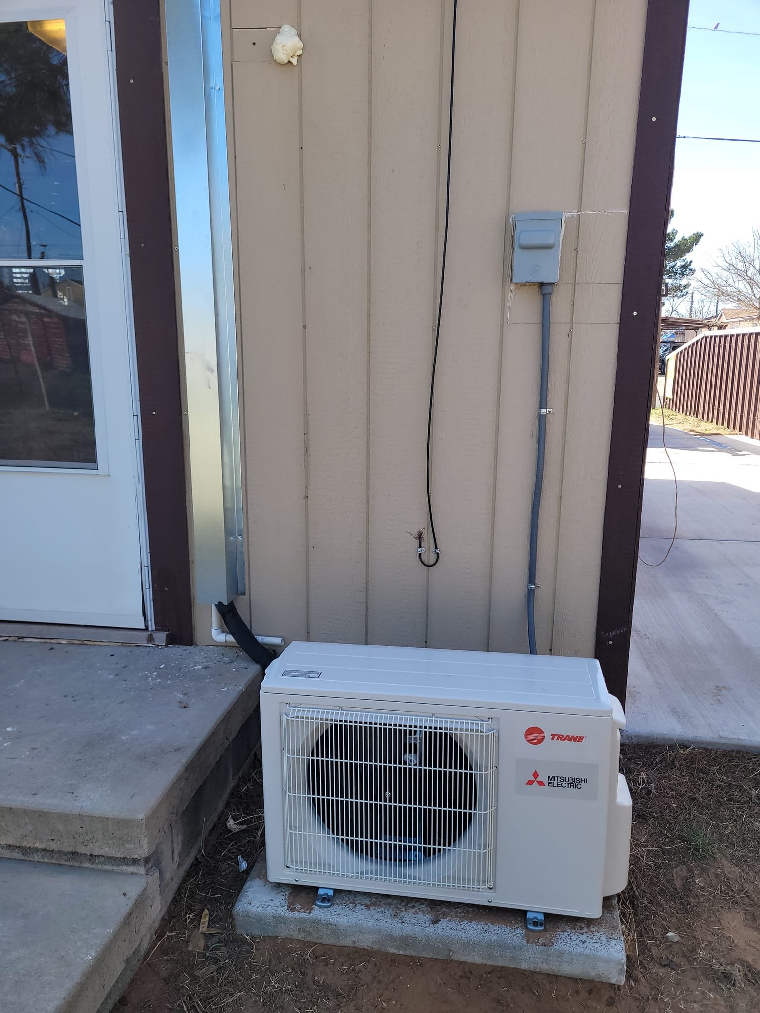 Outdoor air conditioning unit installed next to house wall, with wires and conduit running along the wall.