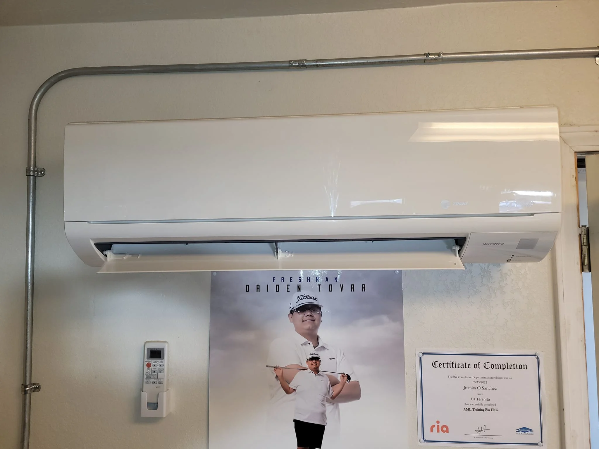 Indoor wall with a large white air conditioner unit mounted near the ceiling. Below it, there is a poster of a young male golfer holding a golf club, wearing a white shirt, cap, and glasses. A remote control is mounted on the wall next to the poster. To the right of the poster, there is a framed certificate of completion for a golf training program.