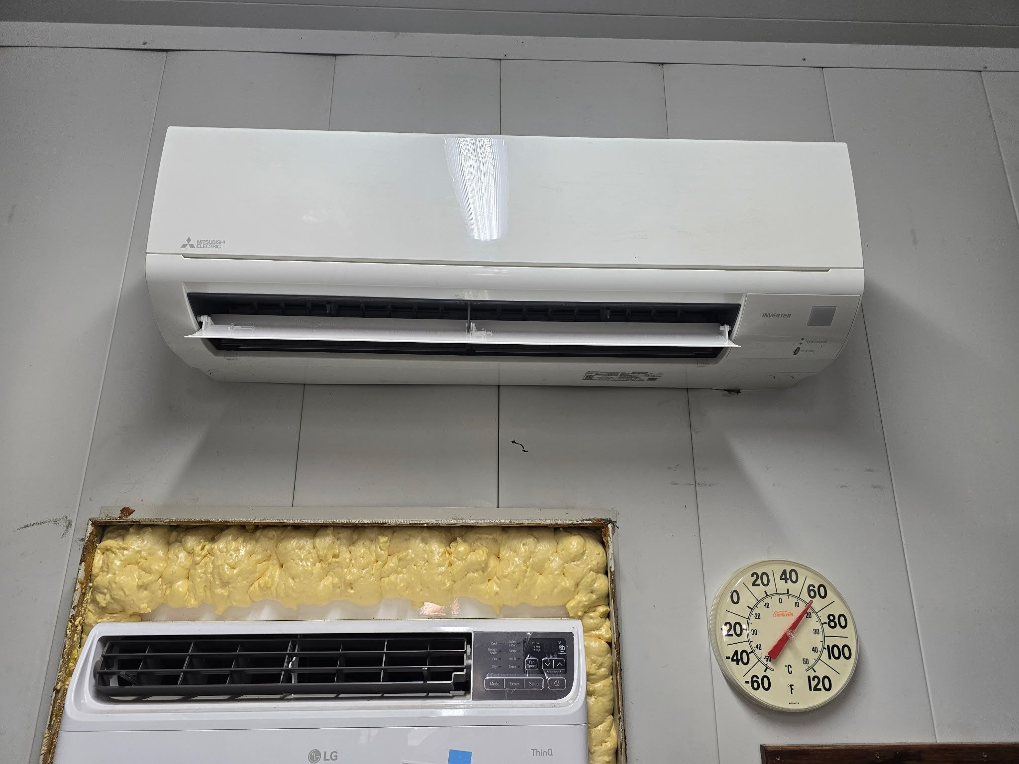 Wall-mounted white Mitsubishi electric air conditioner above a white air purifier with foam insulation around the filter area, a wall clock showing approximately 58°F, and a gray tiled wall.