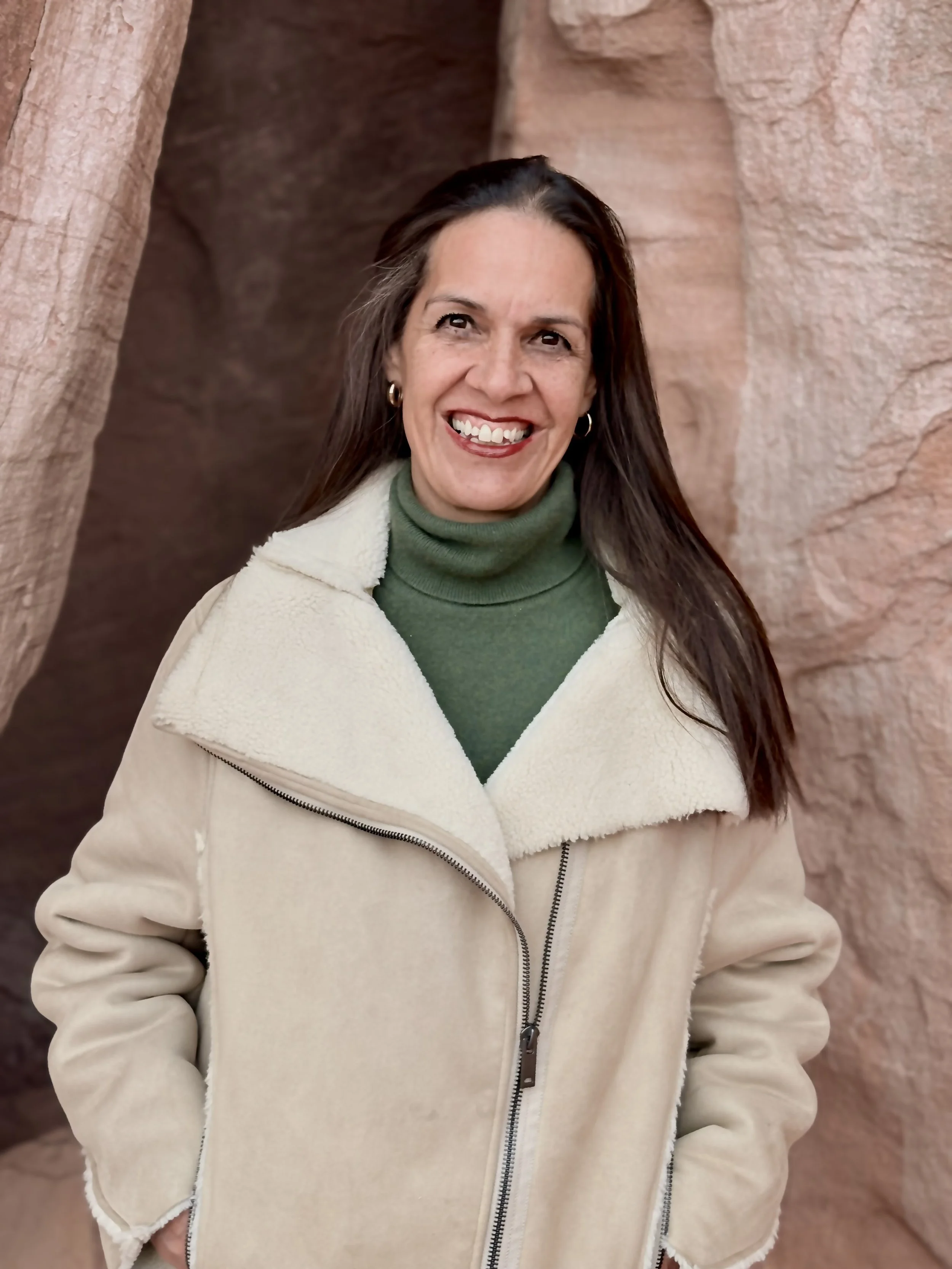 A young woman with brown hair, glasses, and a nose piercing, smiling, standing outdoors against a rocky background, wearing a red floral long-sleeve shirt, with her arms crossed.