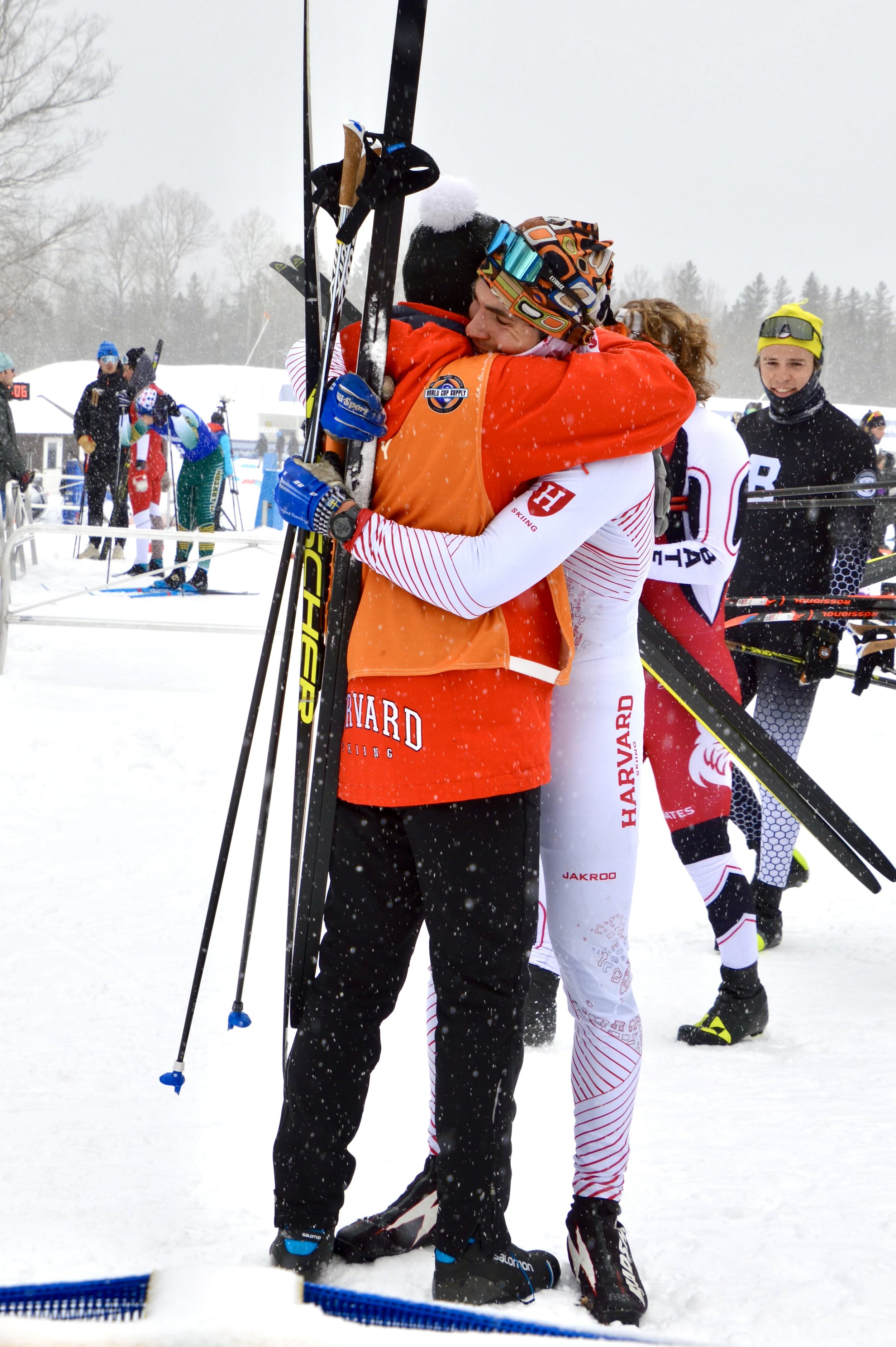 Two cross-country skiers in winter gear hugging each other, one wearing a Harvard outfit, holding skis in a snowy outdoor setting. Other skiers and spectators are visible in the background.