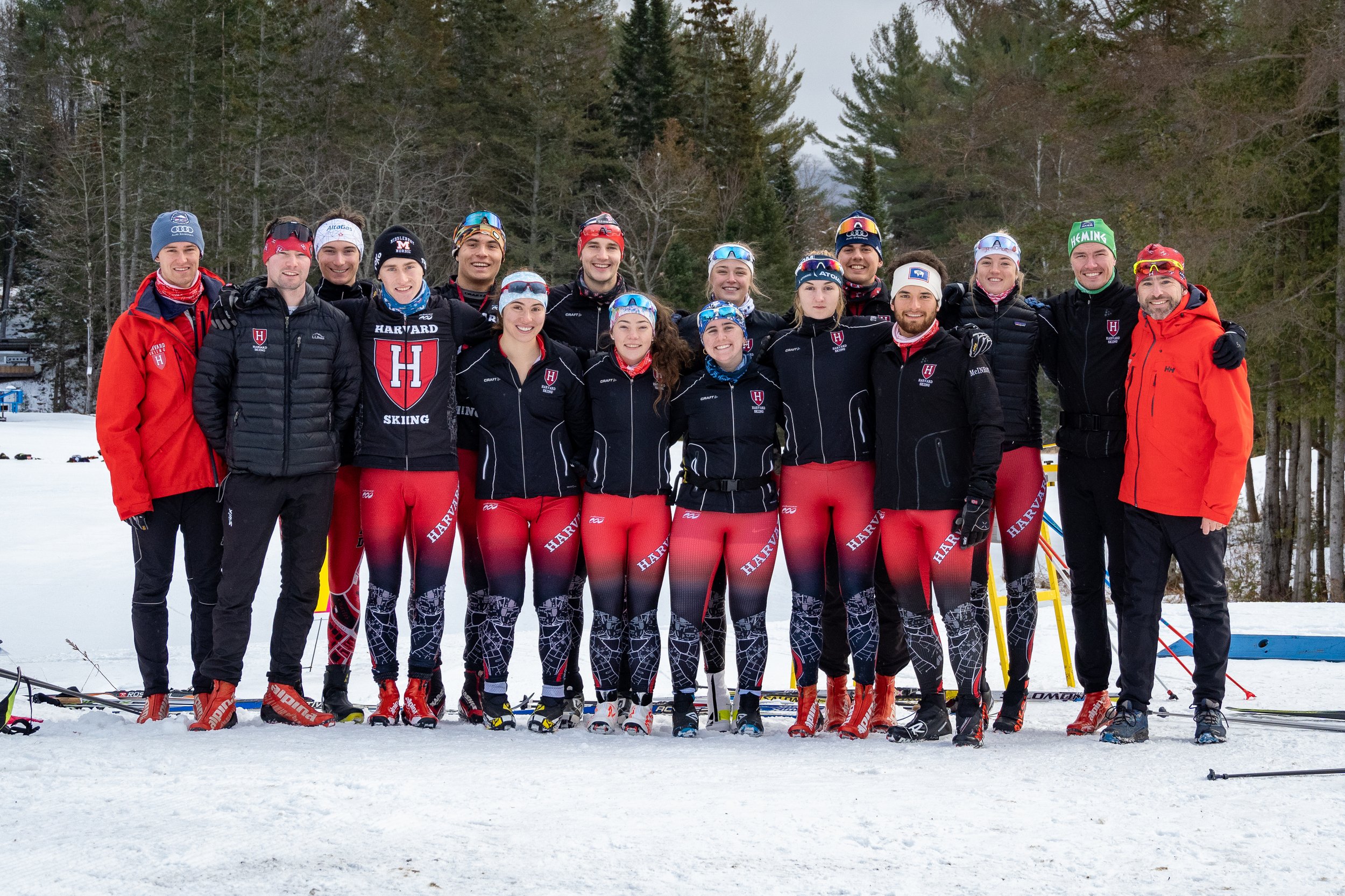 Group photo of a skiing team outdoors, wearing matching red and black outfits, on a snowy landscape with trees in the background.