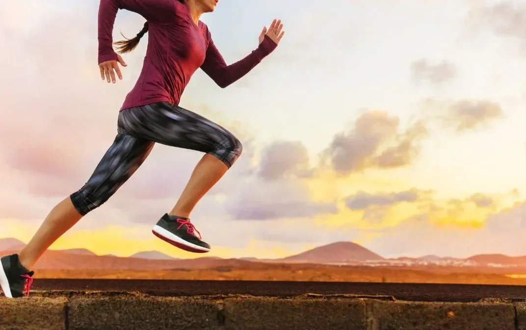 A young woman in athletic attire running outdoors during sunrise or sunset, with mountains in the background.
