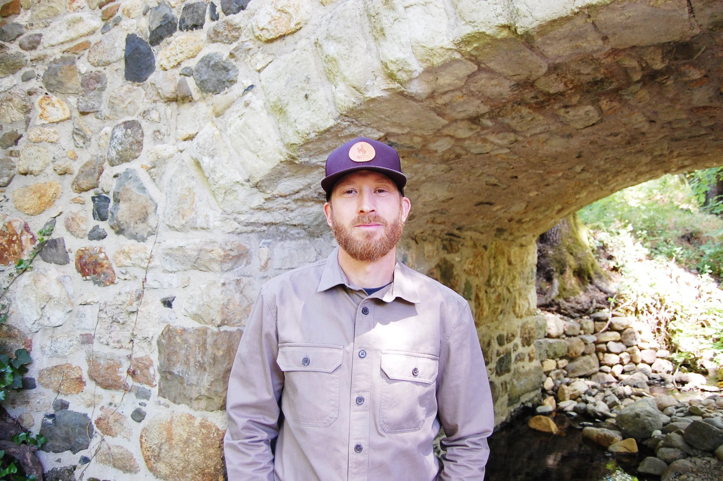 A man with a beard stands outdoors among trees and bushes, wearing a gray button-up shirt and a black baseball cap with a brown emblem.