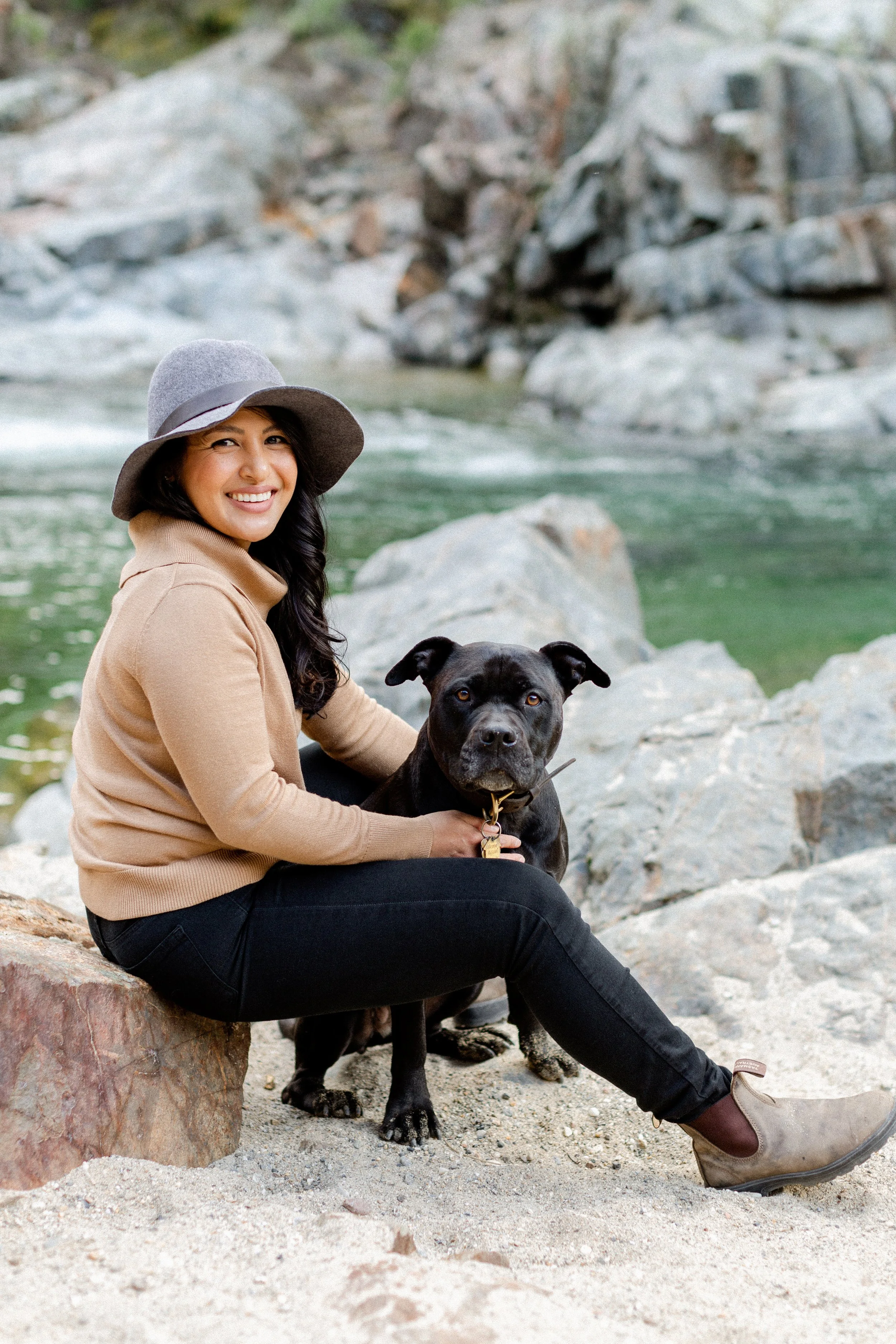 A woman with long black hair wearing a khaki button-up shirt, smiling, standing outdoors in a sunlit green forest with trees and foliage.