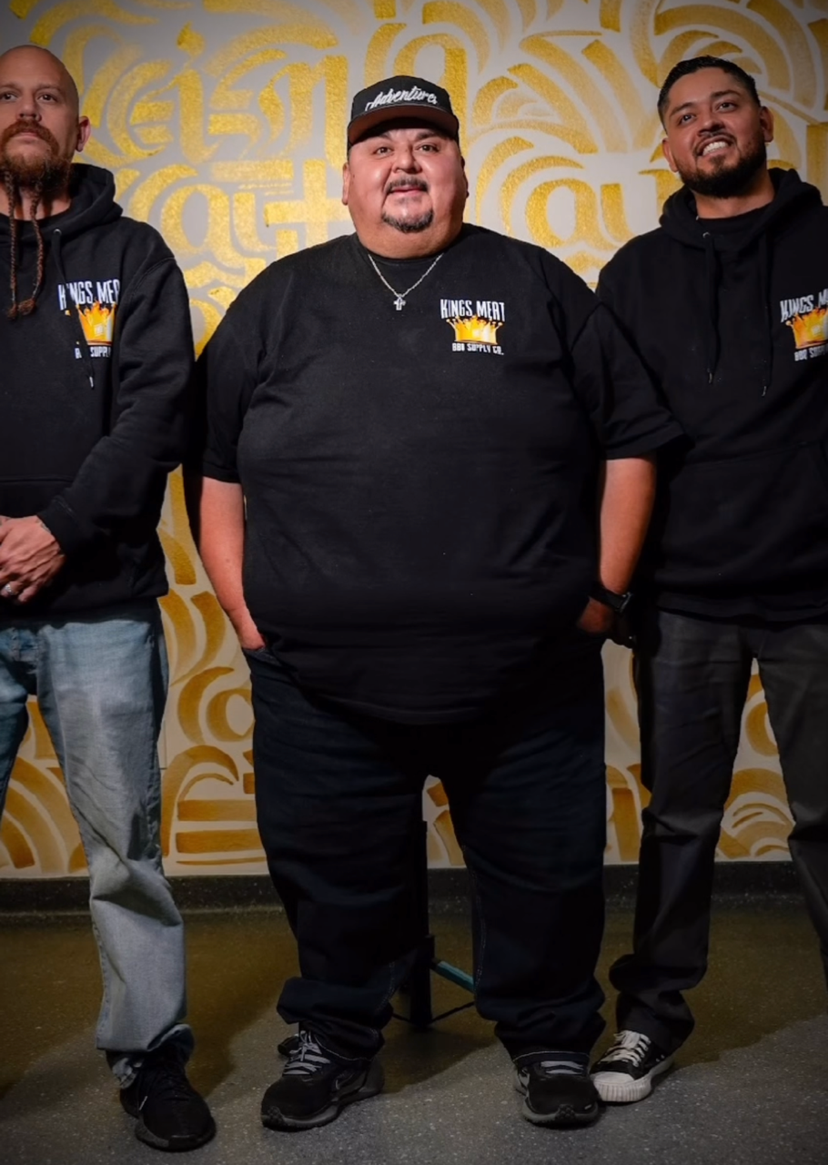 Three men standing together against a yellow wall with intricate patterns. All three are wearing black hoodies with a logo that reads 'Kings Meat BBQ Supply Co.' The man in the middle is wearing a black cap and has a featureful build. The man on the right has a beard and is smiling, while the man on the left has long hair and a beard.