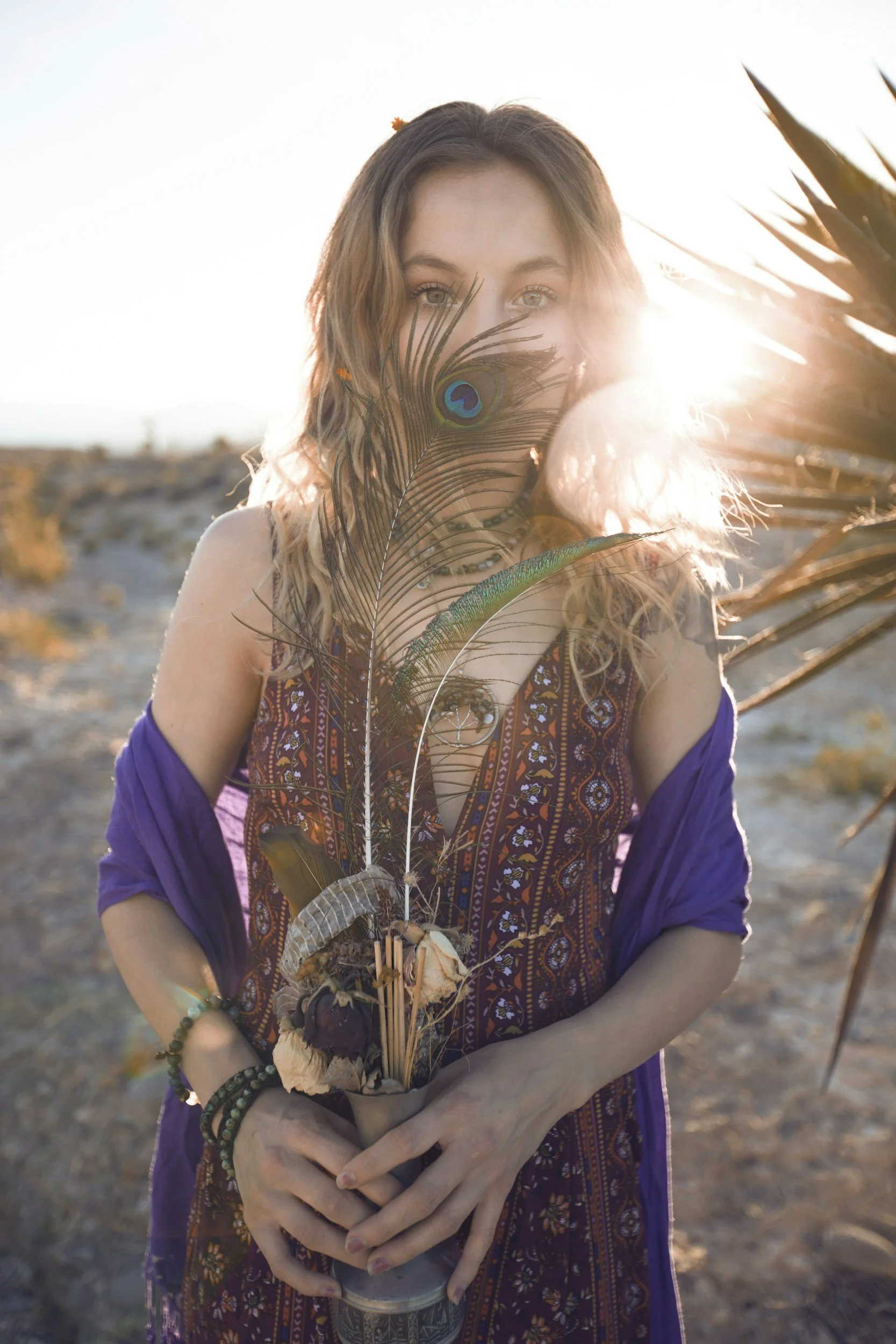 A young woman holding a bouquet of dried flowers and peacock feathers, standing outdoors during sunset with sunlight backlighting her face.