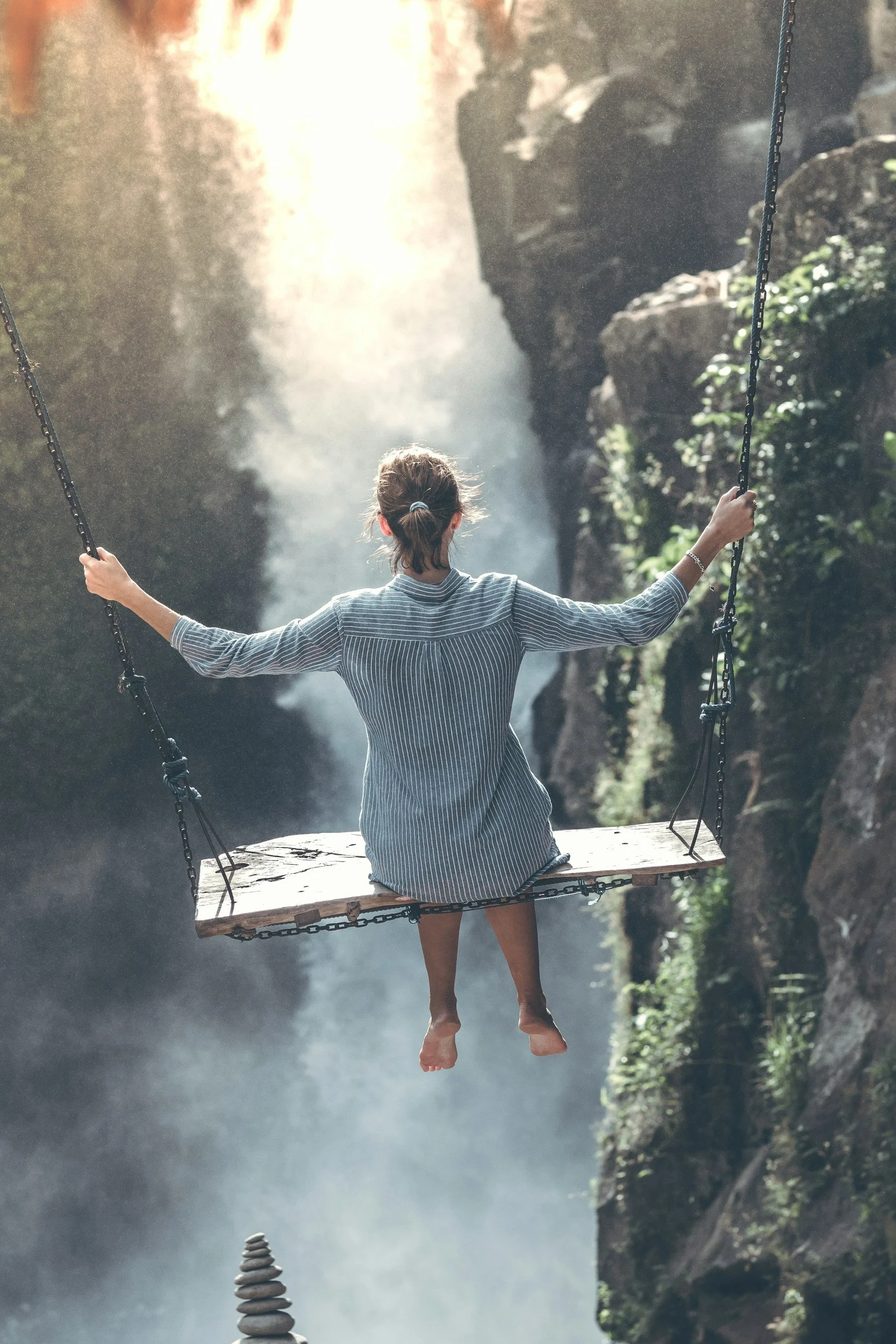 A person with a ponytail and striped shirt is sitting on a wooden swing hanging over a waterfall, with their back to the camera, surrounded by lush greenery and rocks.