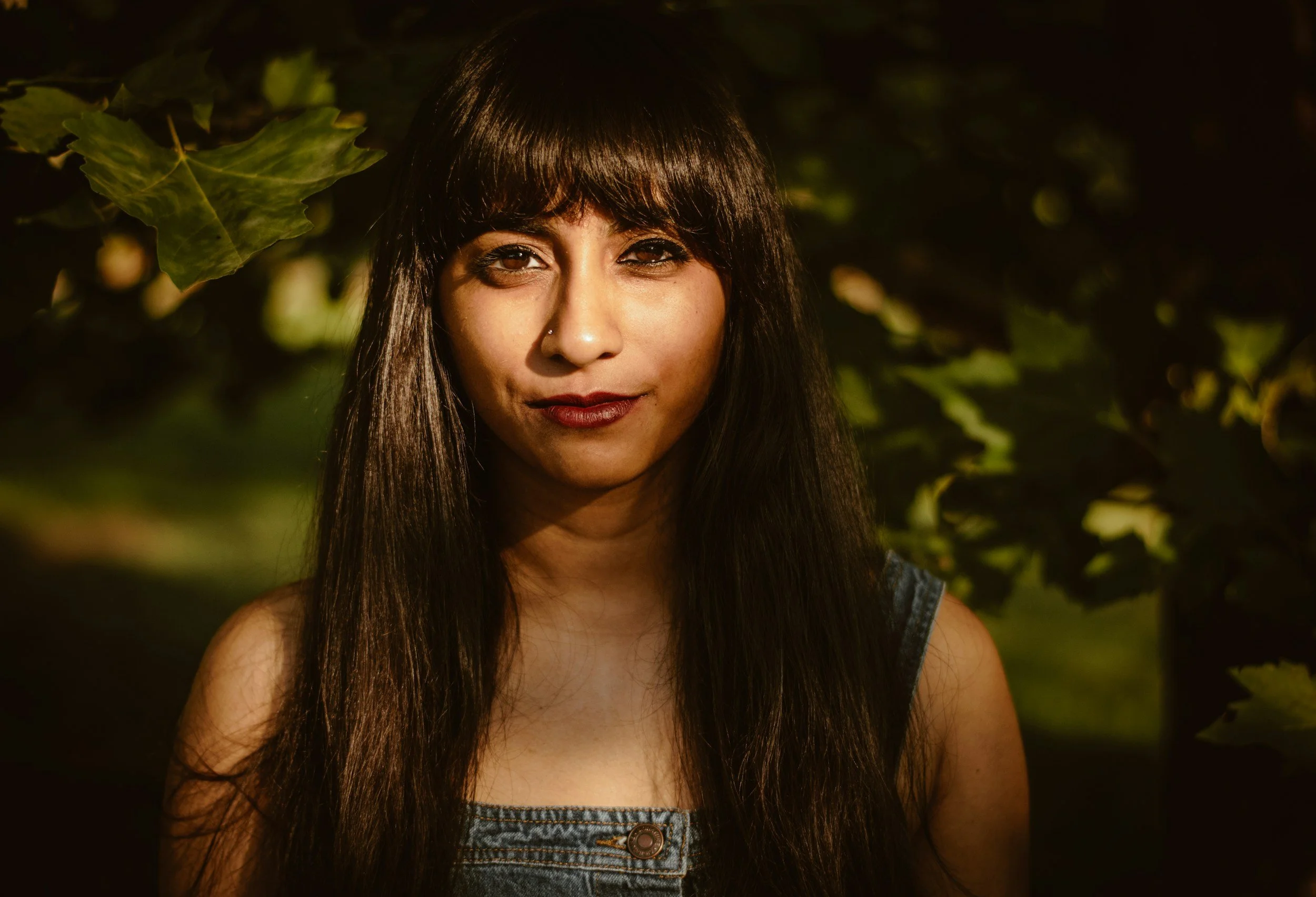 A woman with long dark hair and bangs, wearing a denim top, standing outdoors with green leaves in the background.