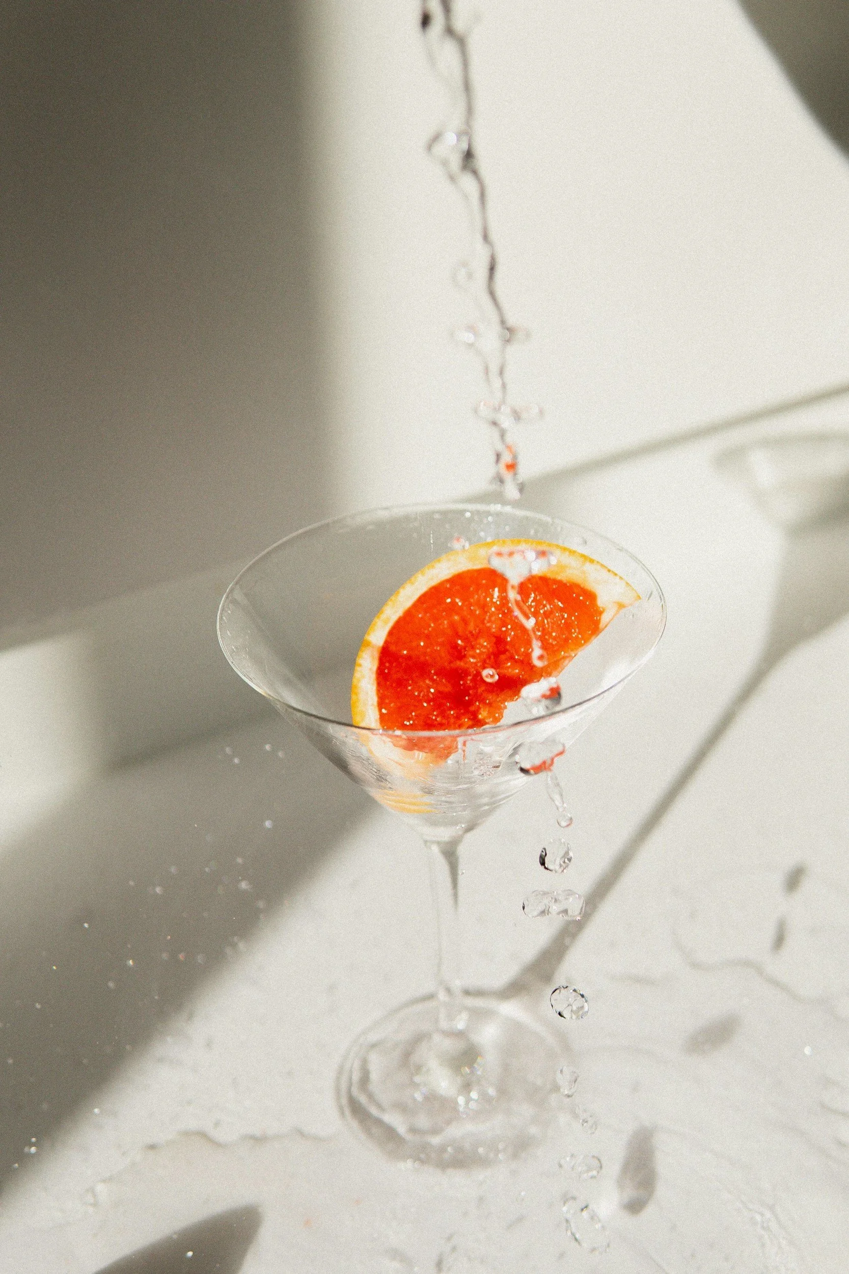 A martini glass with a slice of grapefruit inside, water being poured over it, creating splashes.