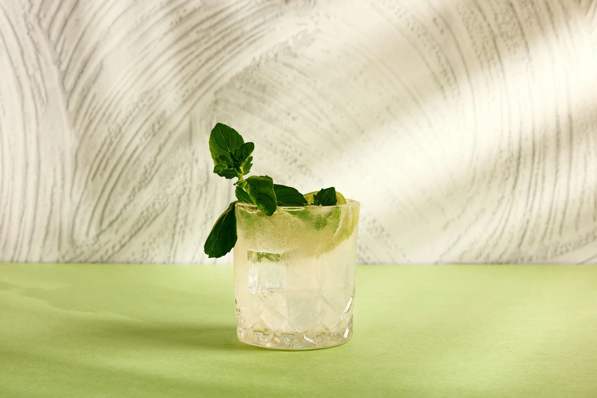 A clear glass containing a light green beverage garnished with fresh mint leaves, placed on a green surface with a textured, light-colored background.