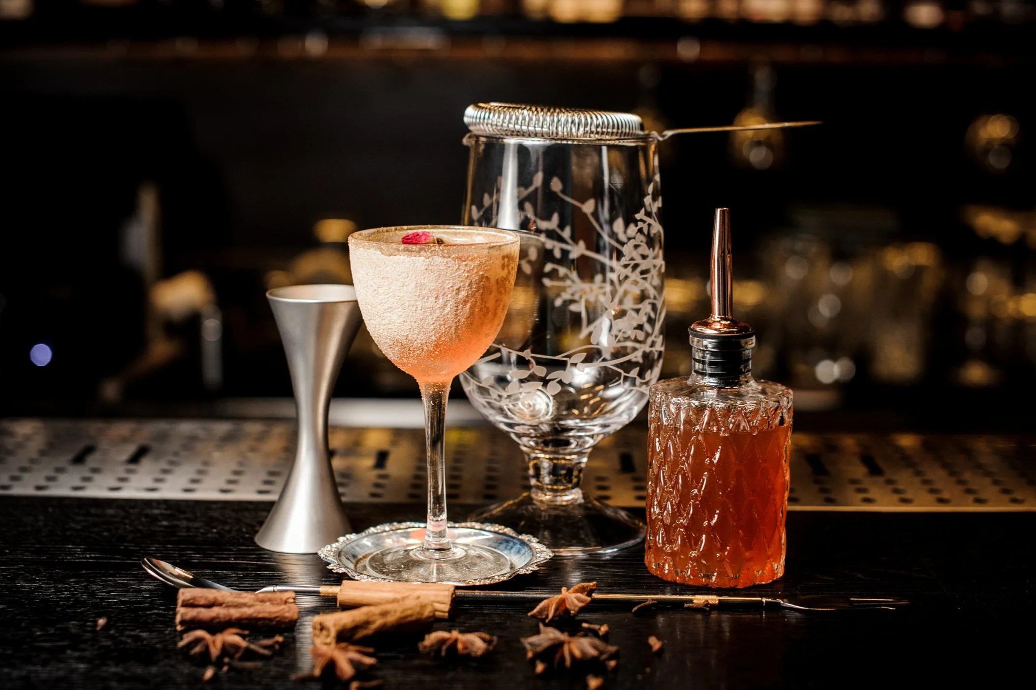Set of barware and cocktails with star anise and cinnamon sticks on a bar counter under warm lighting.
