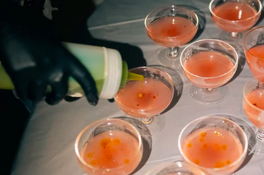 A person wearing a black glove is pouring a pink cocktail into a glass, with several other glasses filled with similar pink drinks on the table.