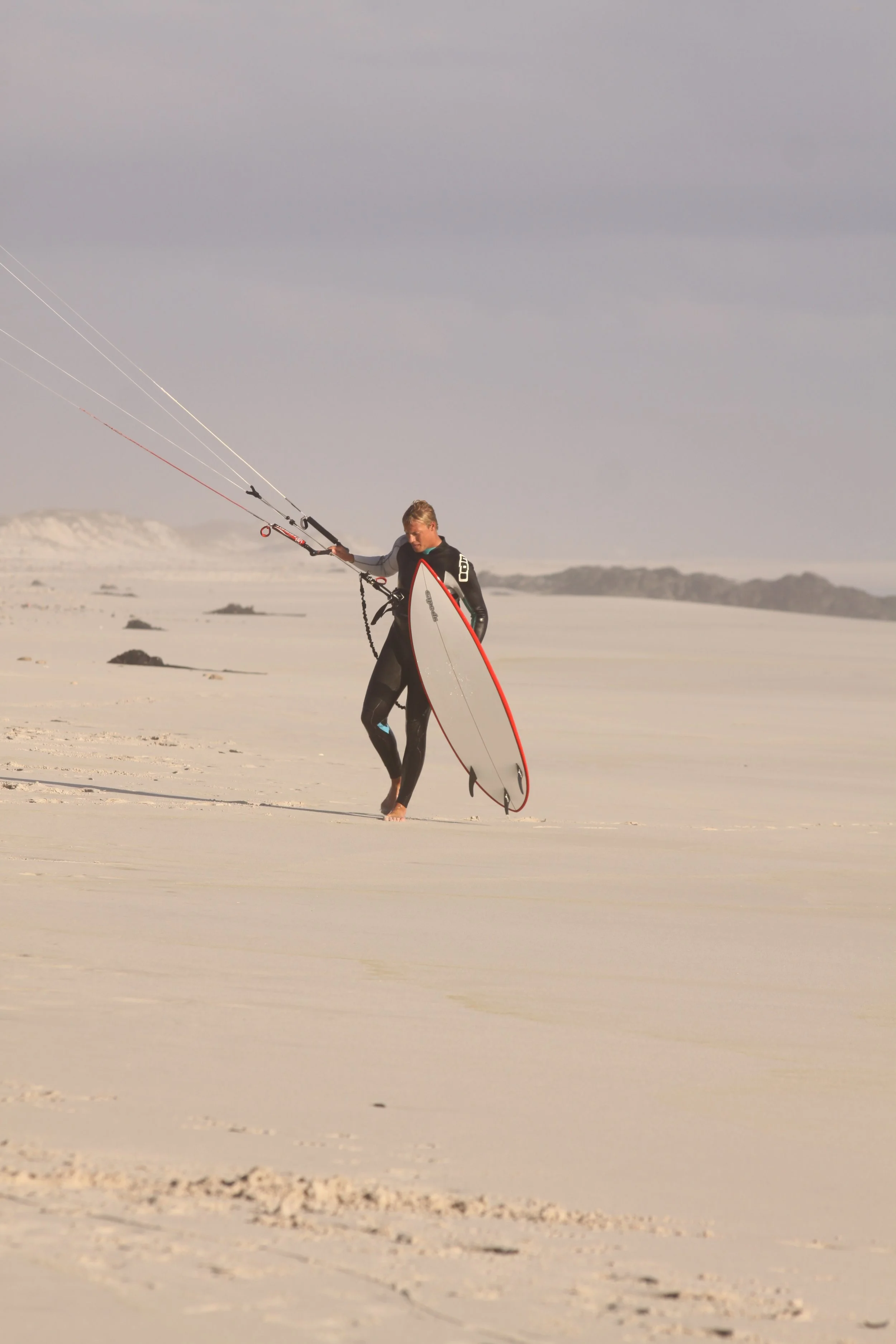 Ein Mensch trägt einen Surferanzug und hält eine große Surfbretthalterung, während er barfuß auf einem weißen Sandstrand mit Kite-Ausrüstung steht.