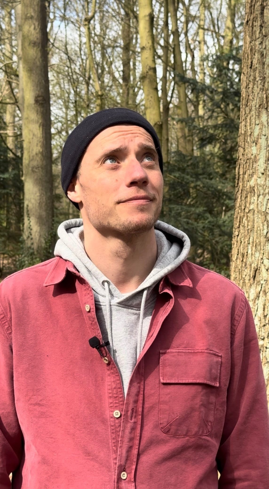 Ein junger Mann in einem Wald blickt nach oben, trägt eine schwarze Mütze, einen grauen Hoodie und eine rotes Hemd.
