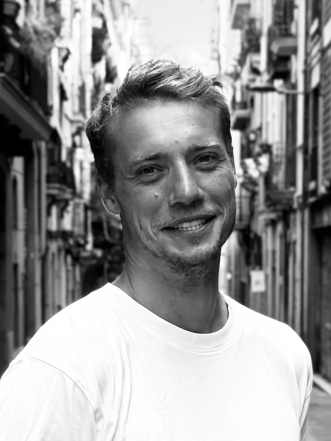 Junger Mann mit hellen Haaren, smiling, trägt ein weißes T-Shirt, steht auf einer engen Straße in einer Stadt, im Hintergrund alte Gebäude.