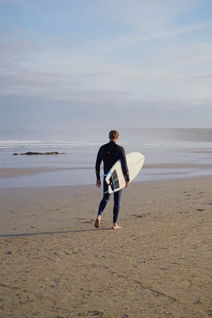 Ein Mann in Neoprenanzug, der barfuß am Strand läuft, ein Surfbrett unter dem Arm tragend, Blick aufs Meer bei bewölktem Himmel.