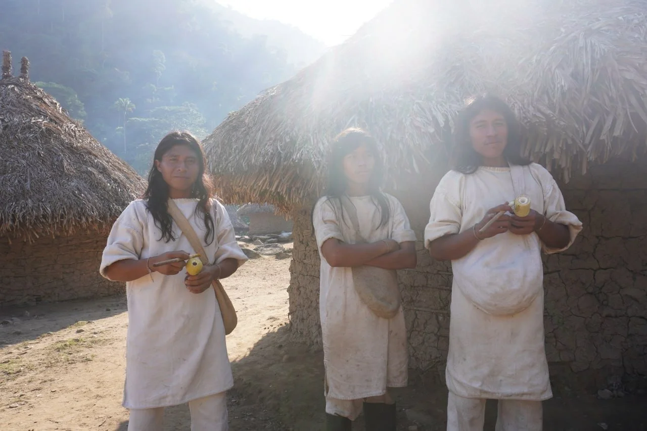 Drei indigene Frauen in weißen Kleidern stehen vor traditionellen Lehmhütten mit Strohdächern, umgeben von einer bewaldeten Berglandschaft bei Sonnenlicht.