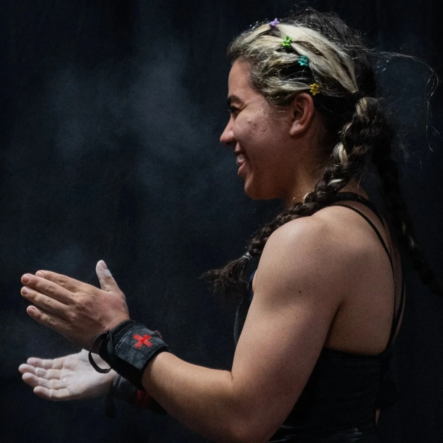 Side profile of a smiling woman with braided hair decorated with colorful clips, wearing workout attire and a wristband with a red cross, against a dark background.