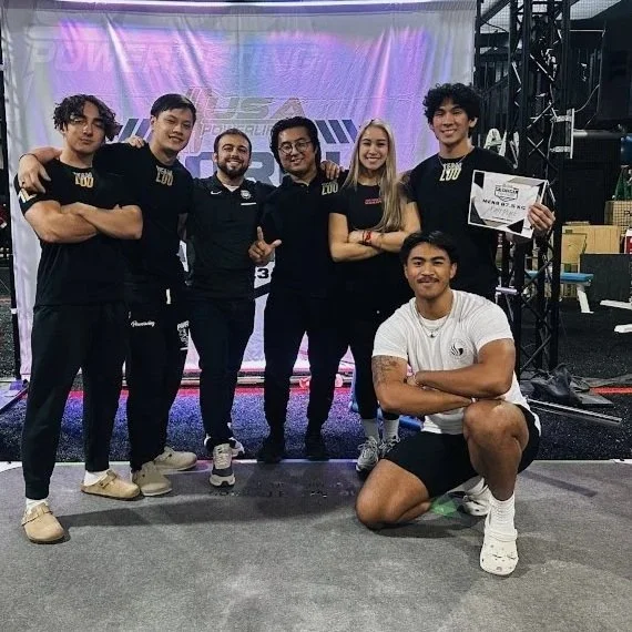 Six young adults standing and one seated man in front, all posing for a photo at a gaming or esports event. The group is smiling, with one person holding a certificate. The background features a stage with a screen displaying the words 'Lexus' and 'VEX.' The setting appears to be an indoor event space with equipment and boxes visible on the side.