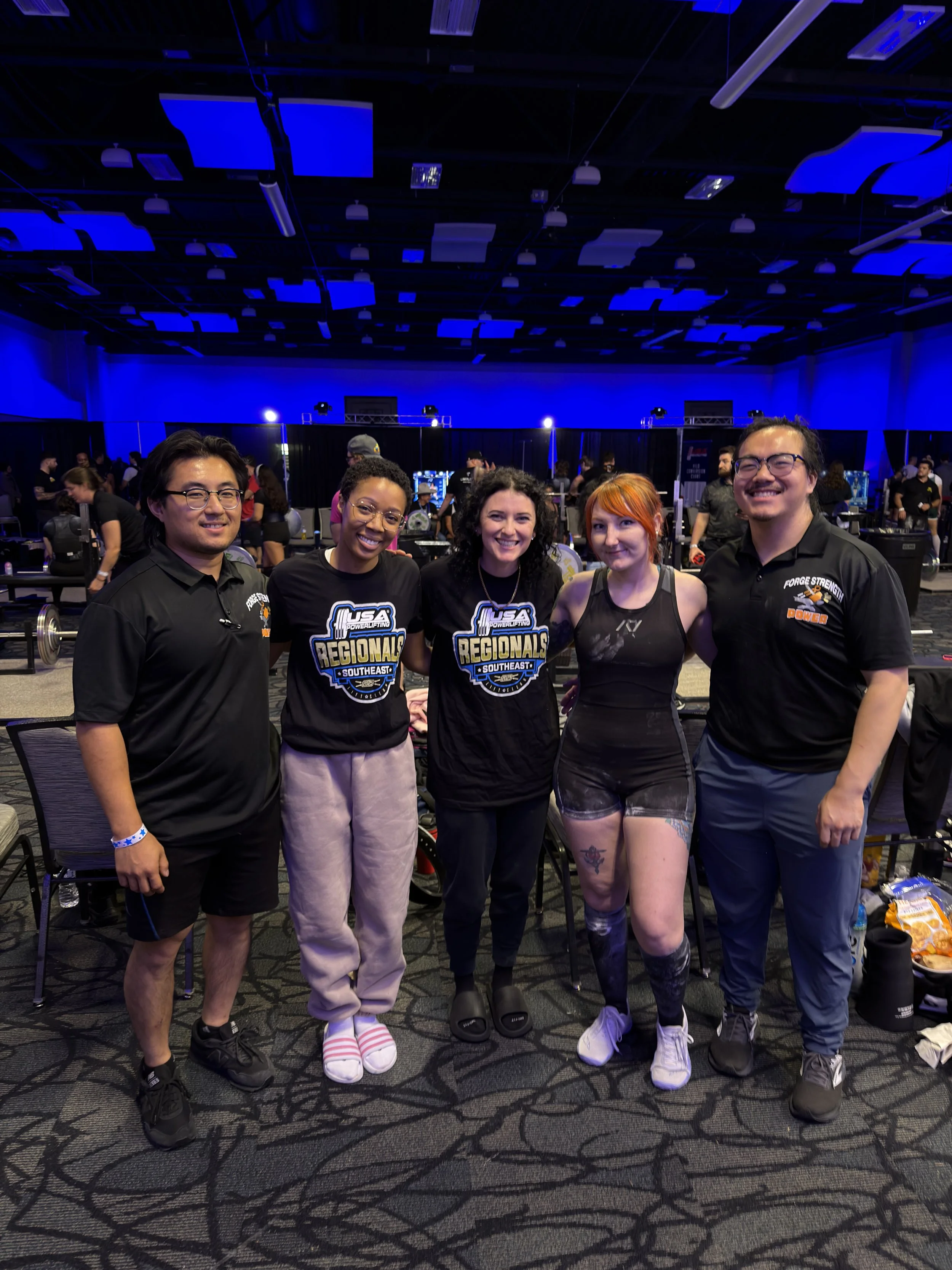 Group of five people, three women and two men, standing together in a dimly lit room with a blue hue, smiling at the camera. Two women are wearing black shirts with 'USA Powerlifting Southeast Regionals' logos, and the woman in the middle has curly black hair. The women on the right is in athletic wear, with visible tattoos on her thighs and arms. The men are in black shirts, one with glasses and the other with a big smile. They appear to be at a weightlifting competition or event.