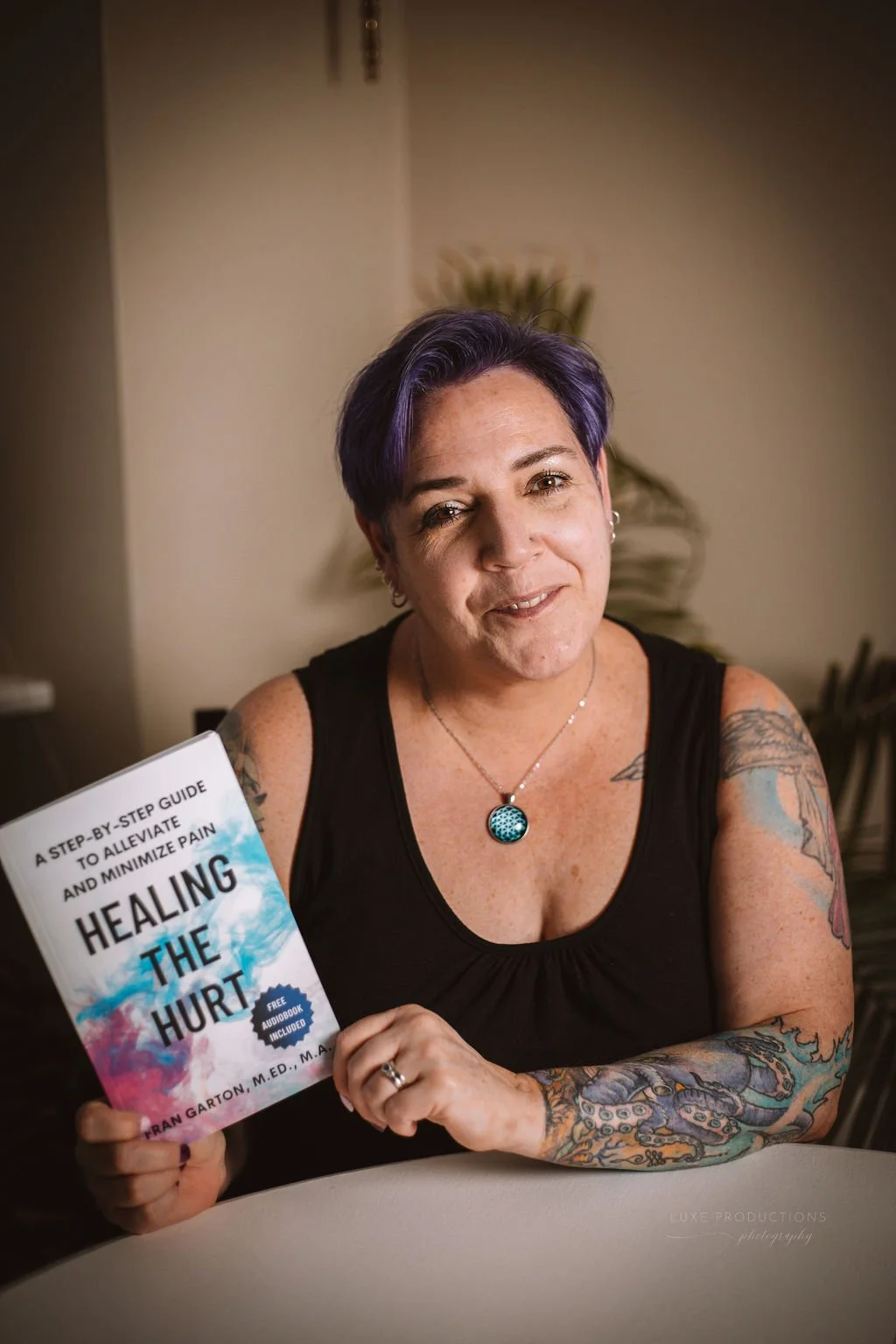 A woman with purple hair and tattoos, wearing a black tank top and jewelry, holding a book titled 'Healing the Hurt' and smiling at the camera.