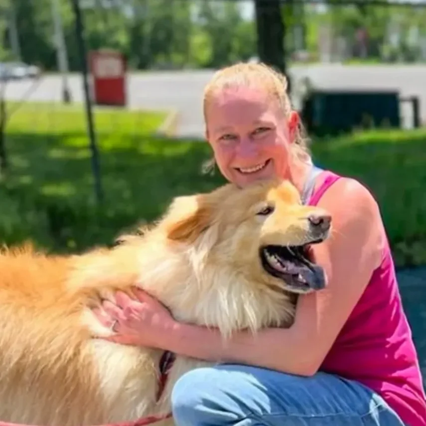 A woman in a pink tank top sitting outdoors, hugging a large, fluffy golden retriever with a happy expression, on a sunny day in a park or grassy area.