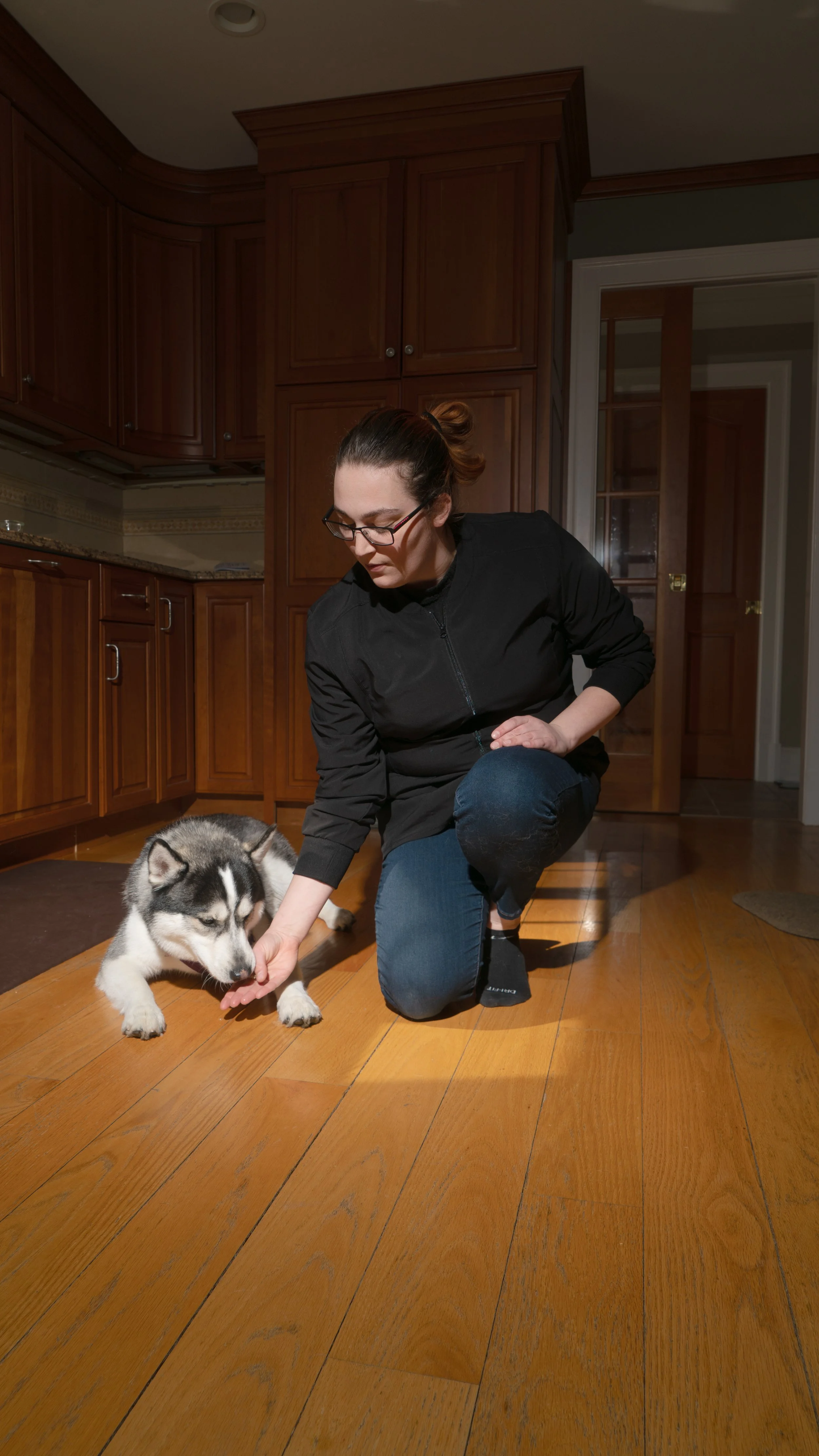 A woman with glasses in a black jacket and blue jeans kneels on a wooden floor, gently touching a Siberian Husky puppy's paw inside a kitchen with wooden cabinets.