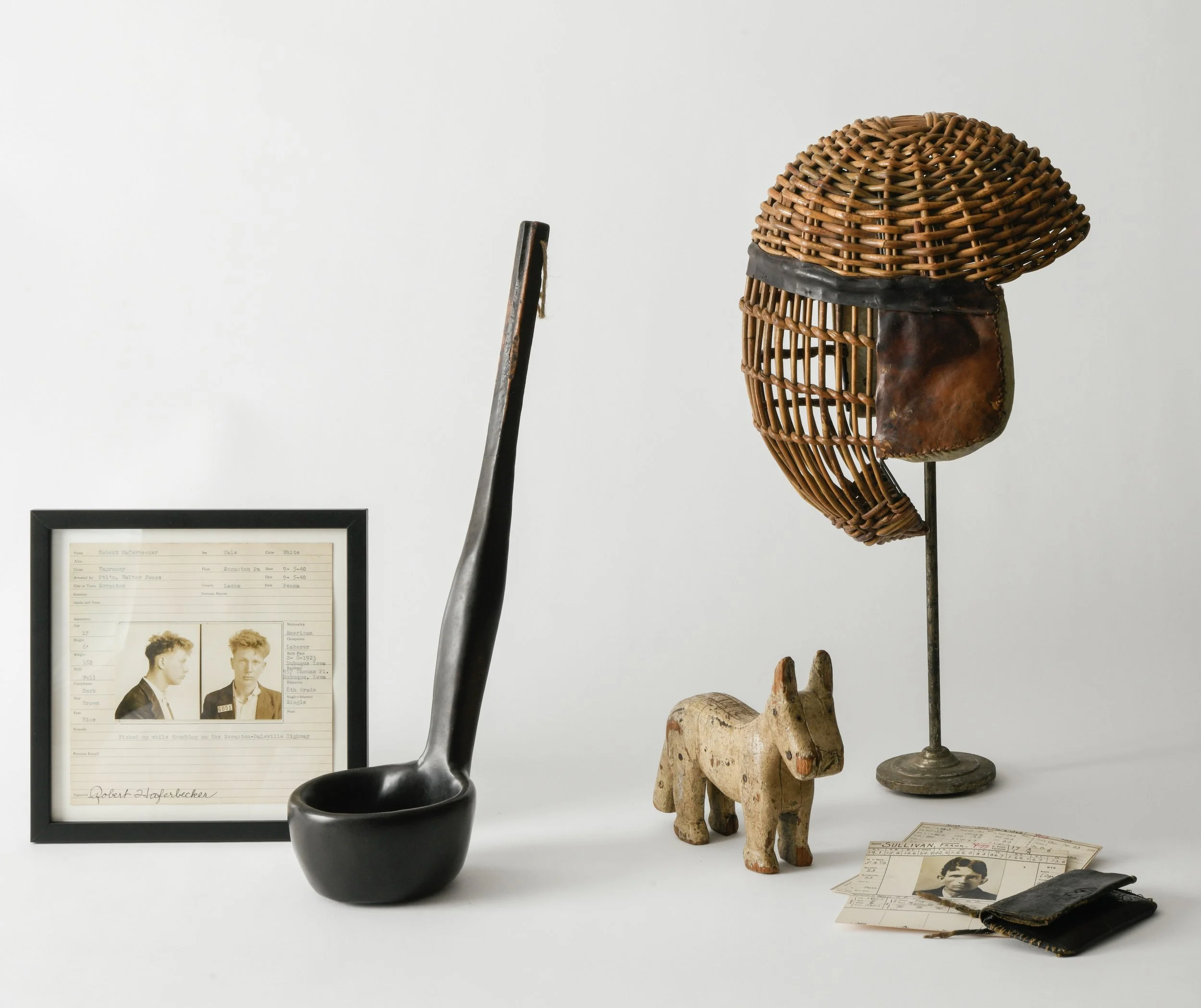 Collection of vintage items including a black ceramic pitcher, a wooden carved donkey figure, a woven wicker sculpture, a framed historical photograph of two young men, and some old documents on a white background.