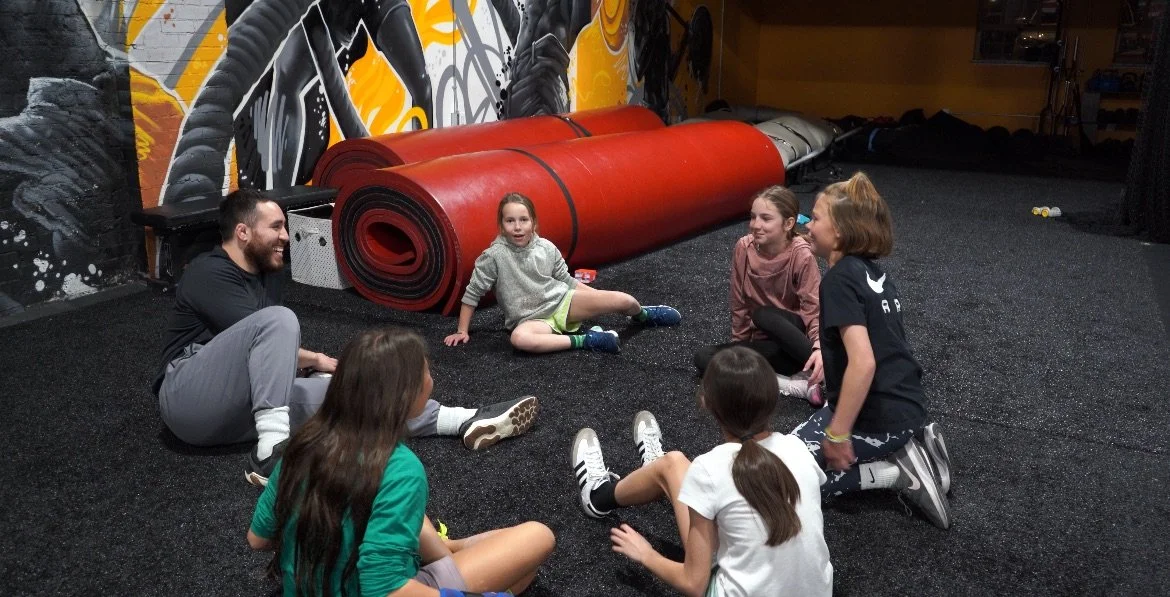 A group of kids and a man sitting in a circle on a black gym floor, laughing and talking, with a red rolled up gym mat and colorful graffiti wall in the background.