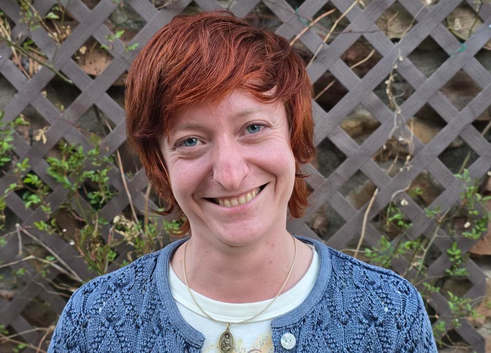 A woman with short red hair and blue eyes smiling in front of a wooden lattice fence with some green plants and dried leaves.