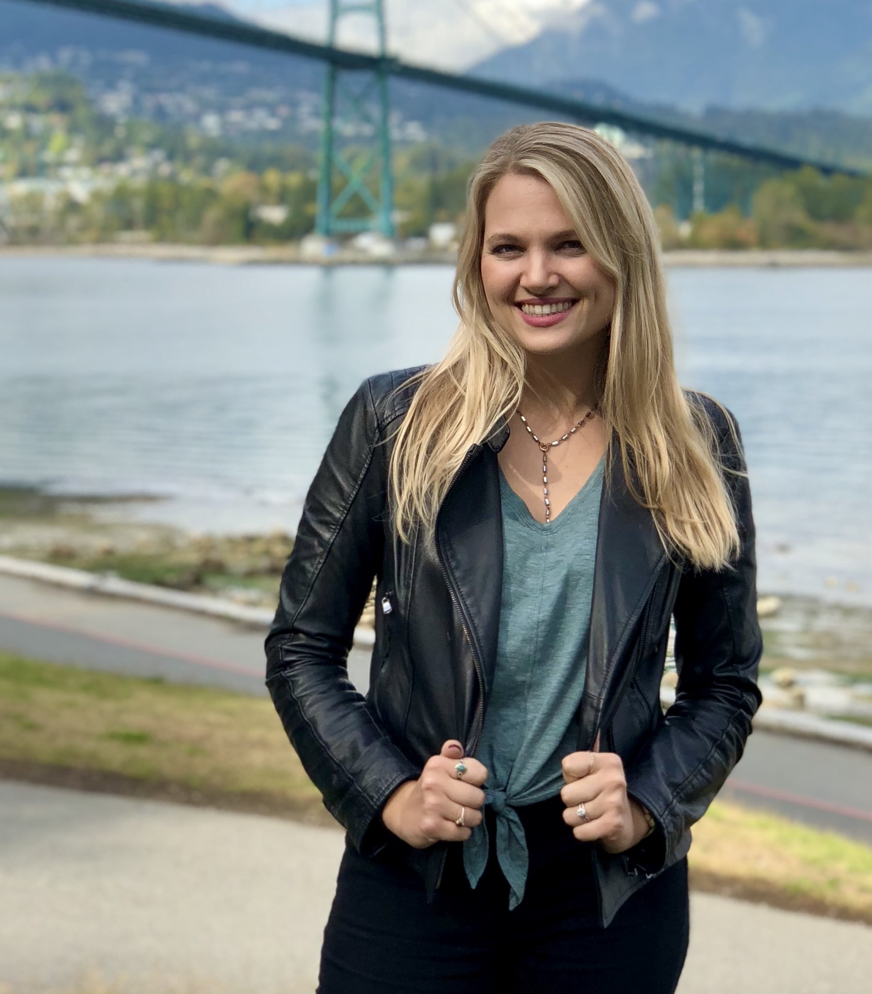 A woman with long blonde hair smiling outdoors by a body of water, with a bridge and mountains in the background.