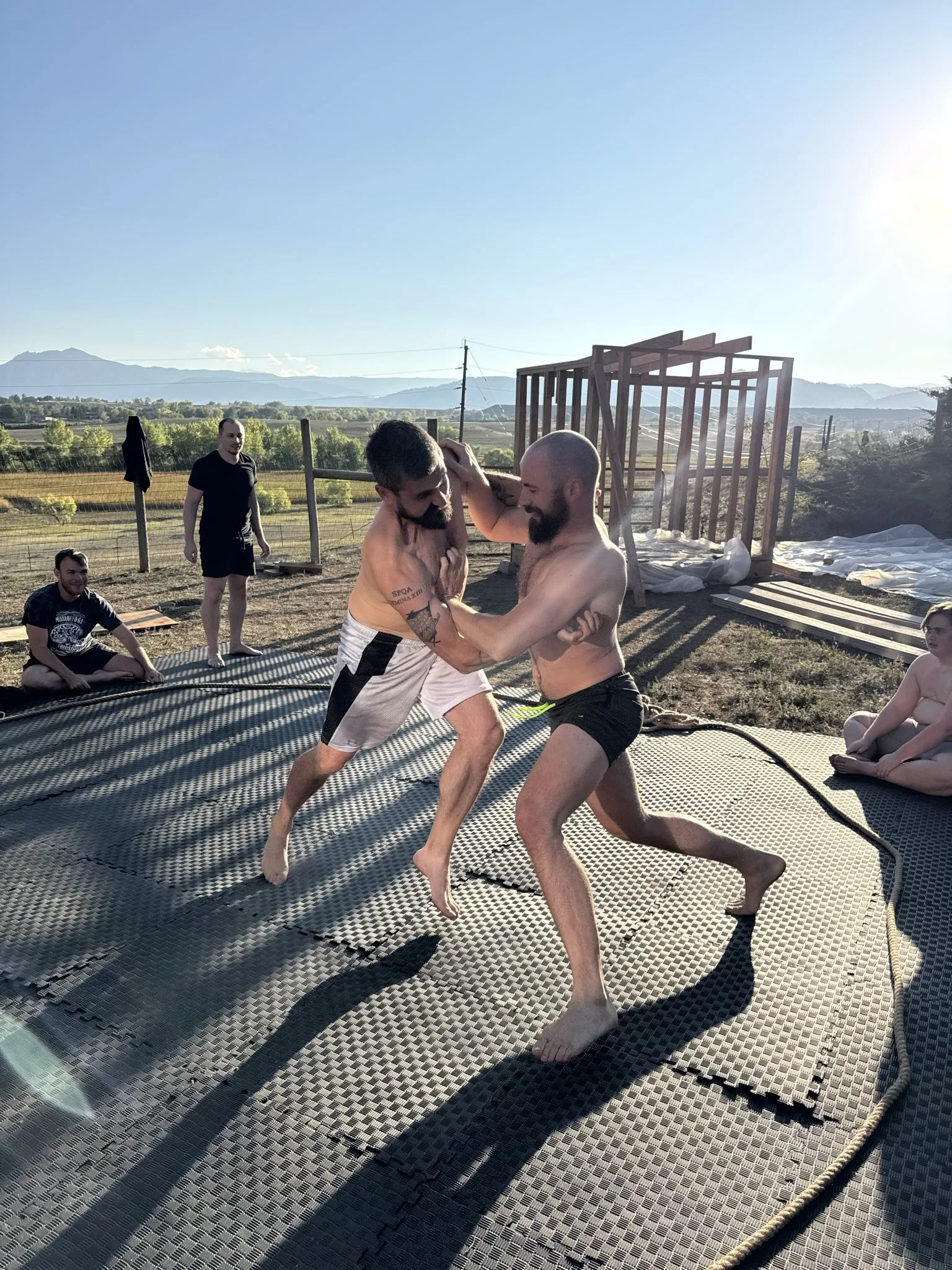 Two men grappling on a trampoline during an outdoor fight, with others watching, in a rural area with mountains in the background.