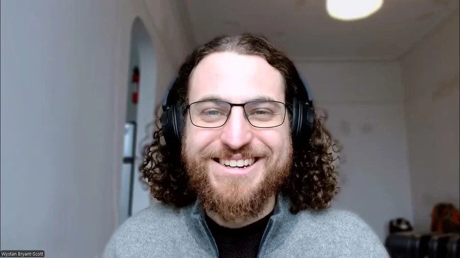 A man with curly hair, glasses, and a beard smiling, wearing headphones and a gray sweater, in a room with a light-colored ceiling and walls.