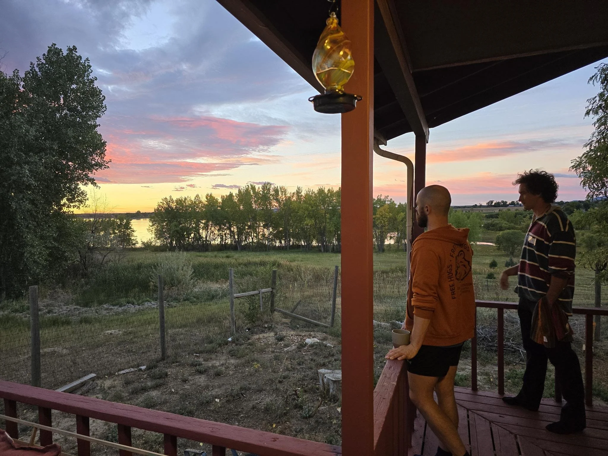 Two people standing on a porch, looking out at a sunset over a river and trees in the distance. The porch has a wooden railing and posts, with a hanging light fixture visible. One person has a shaved head and a beard, holding a cup, and the other has curly hair and is wearing a striped shirt.