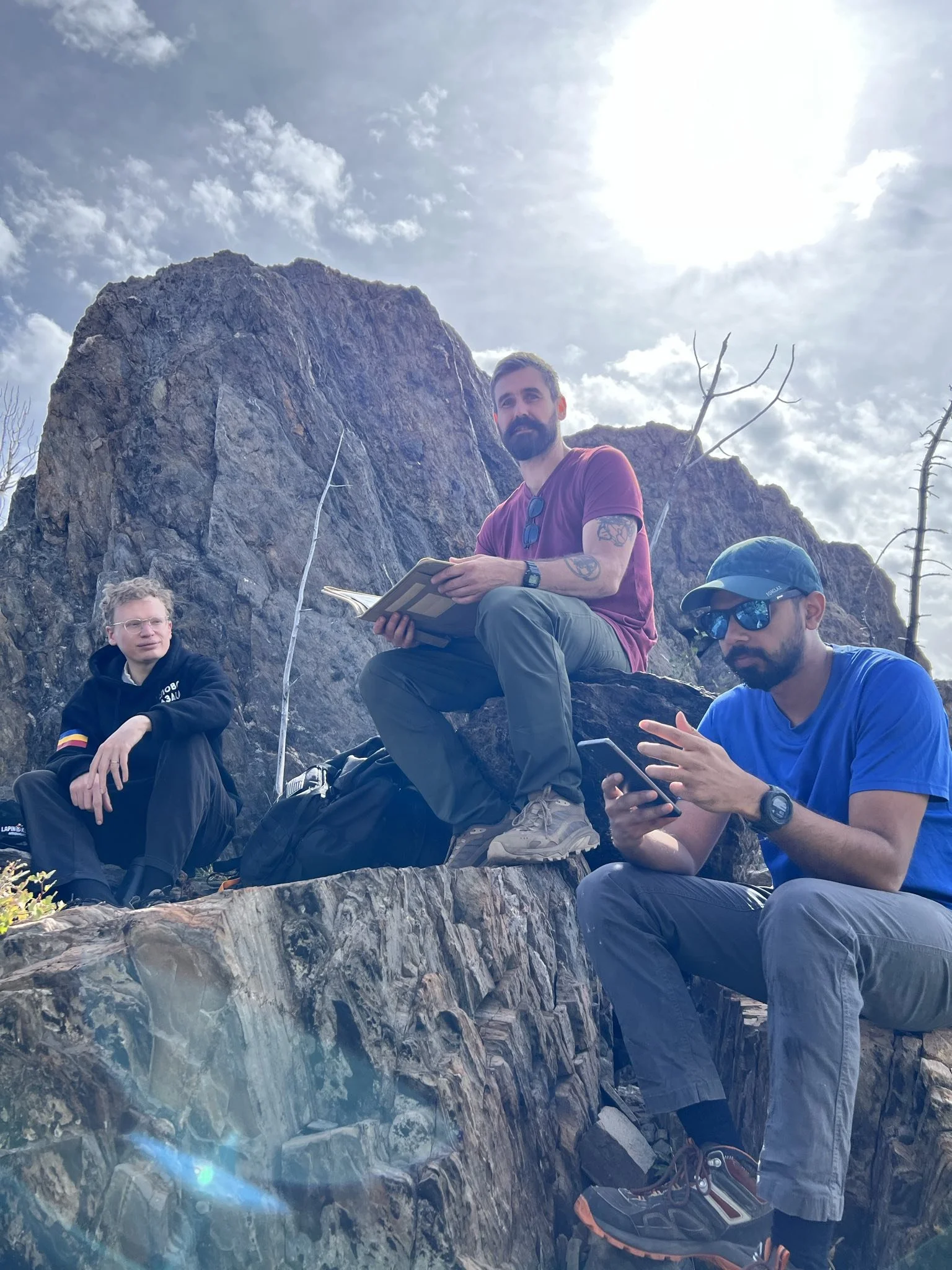 A group of four men sitting on a rocky ledge outdoors, with rocks and a partly cloudy sky in the background. One man is wearing a red shirt and holding a notebook, another is in a blue shirt and sunglasses looking at his phone, a third in a black hoodie and glasses, and the fourth with a beard and tattoos on his arm.