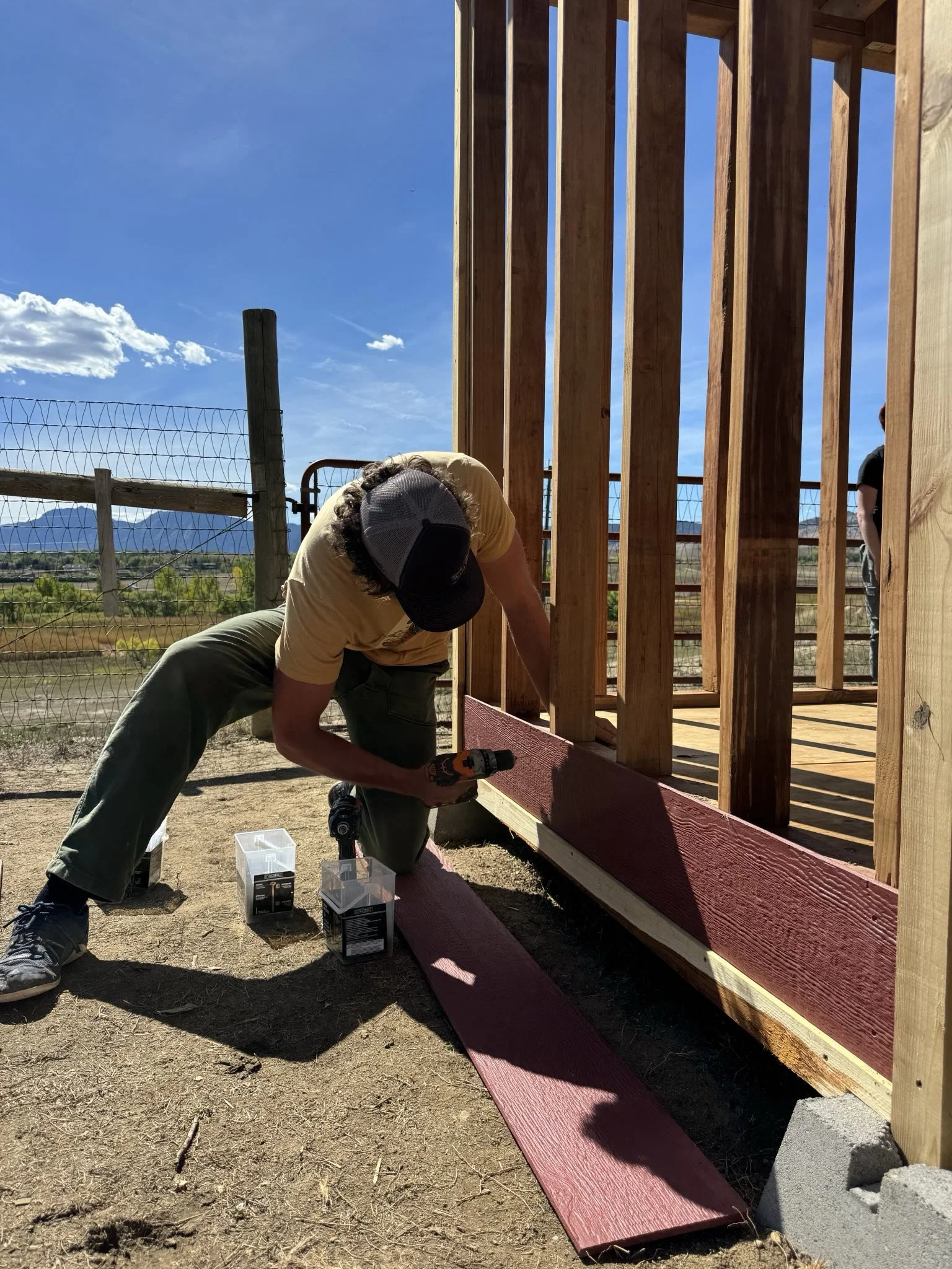 Person working on building a wooden deck outside, wearing a cap and yellow shirt, using a drill to screw in a piece of wood, with red-painted boards on the ground and a wooden railing under construction. Clear blue sky with some clouds in the background.