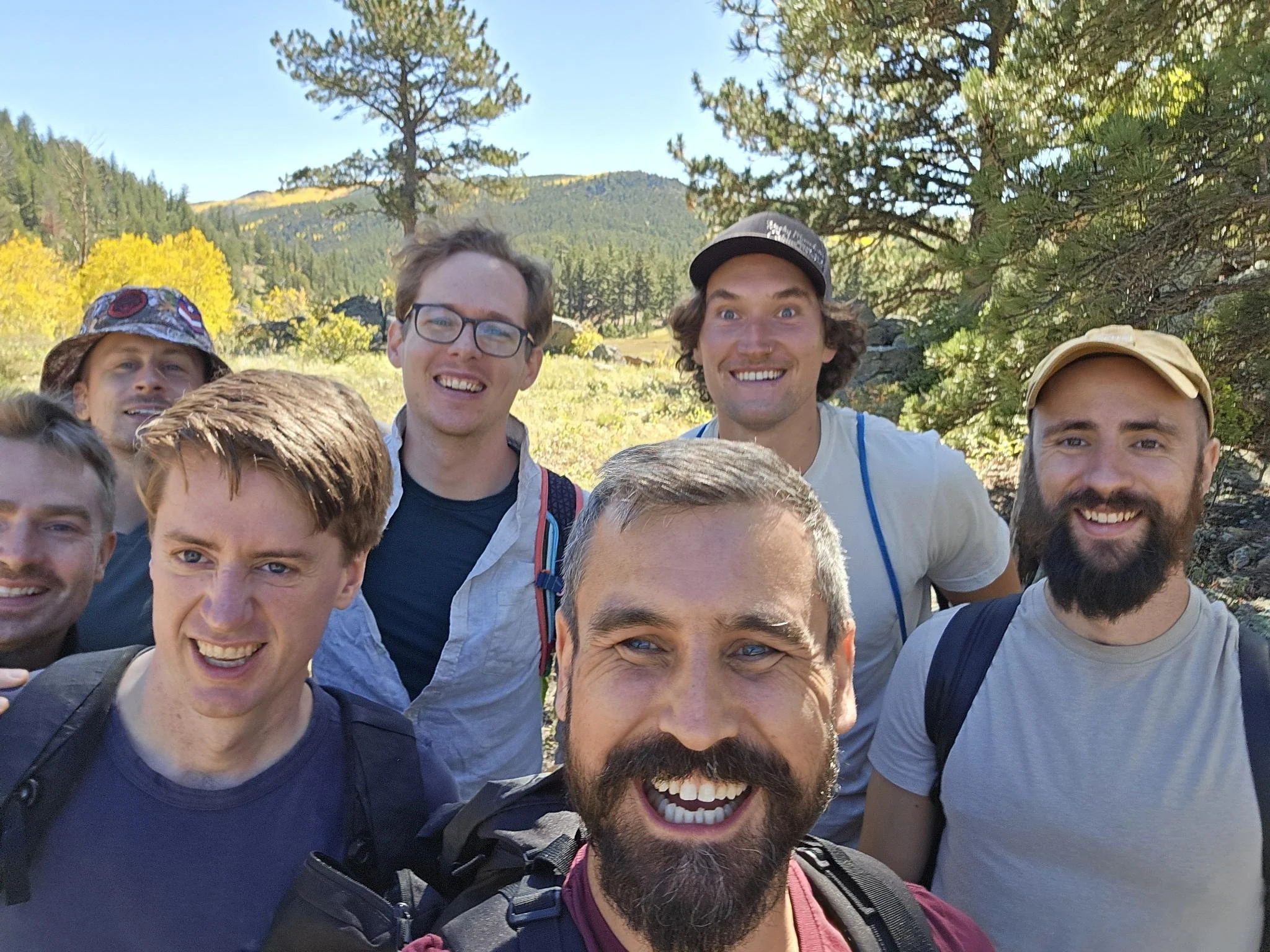 A group of smiling people outdoors in nature, with mountains and trees in the background.