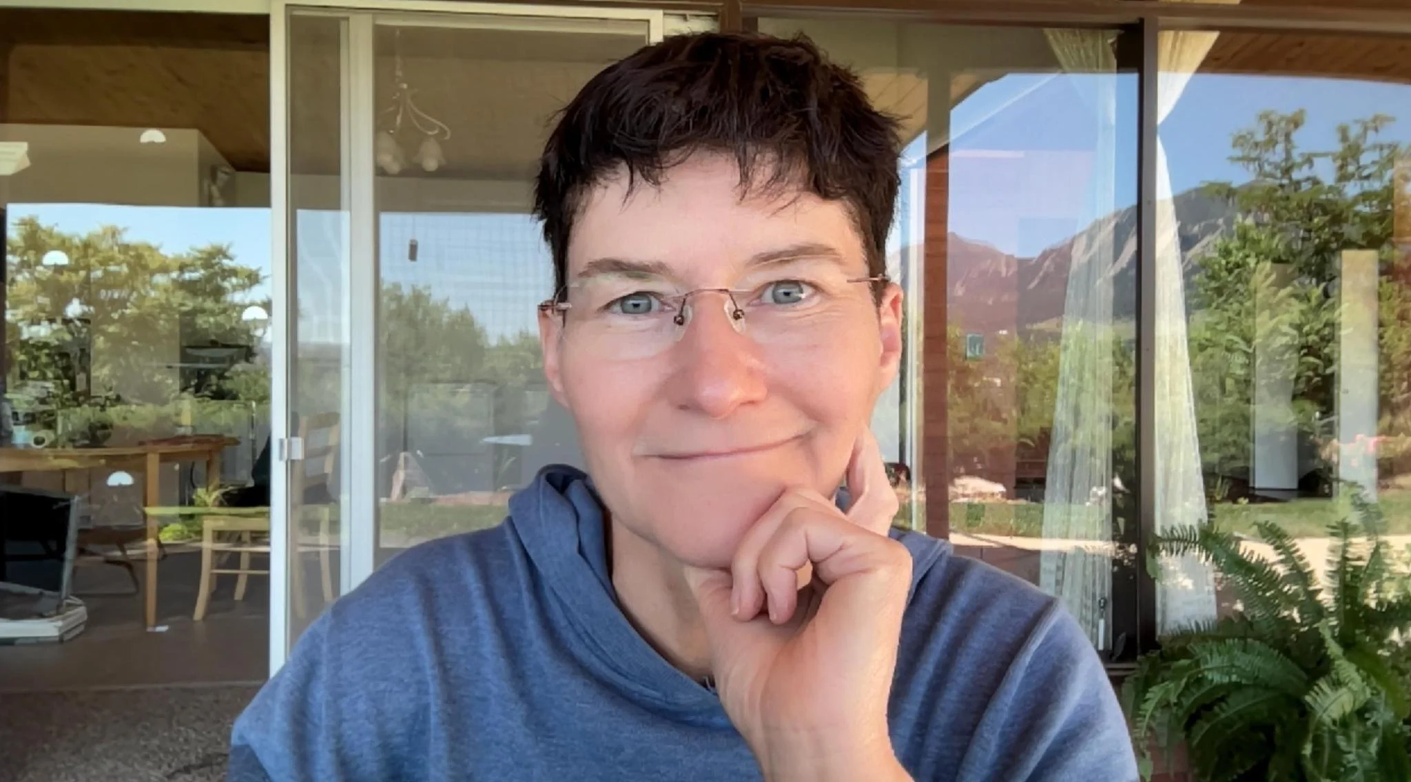 Portrait of a woman with short dark hair, glasses, and a blue hoodie, smiling and resting her chin on her hand in a well-lit room with large windows showing a mountain landscape and greenery outside.
