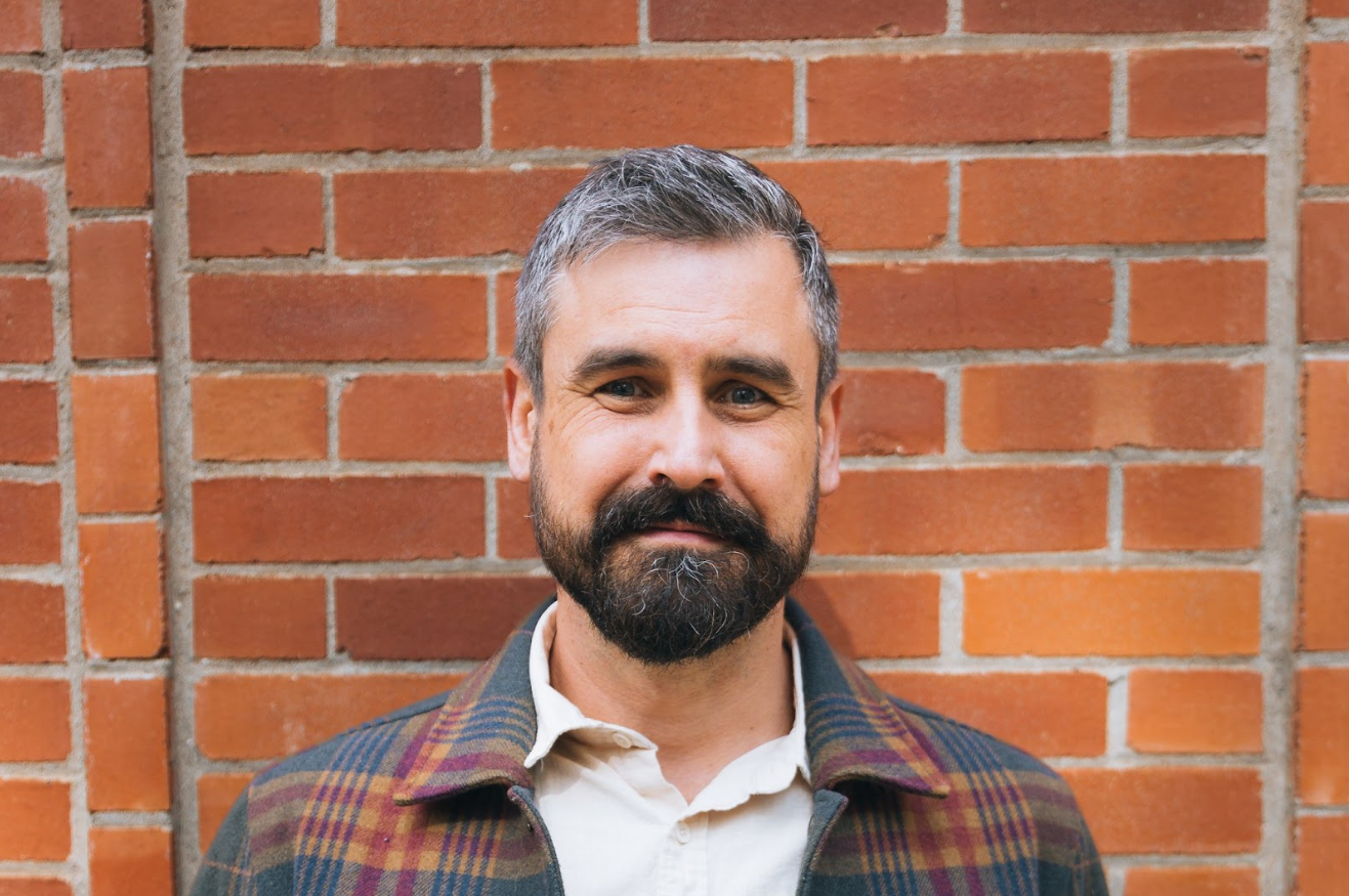 A middle-aged man with short gray hair and a full beard, wearing a plaid jacket over a white shirt, standing in front of a brick wall.