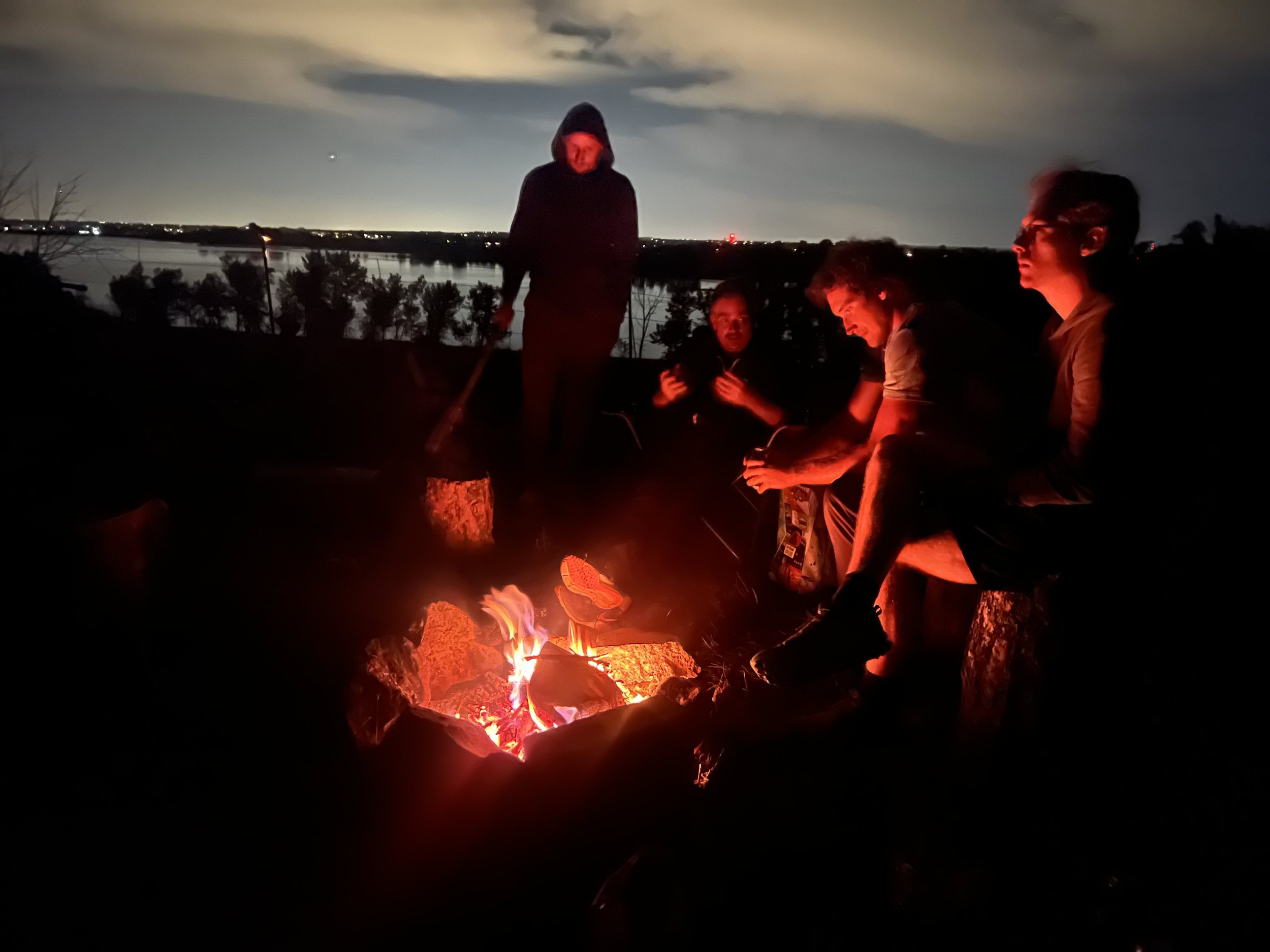 Group of people gathered around a campfire at night, some sitting and one standing, with an overcast sky and a body of water in the background.