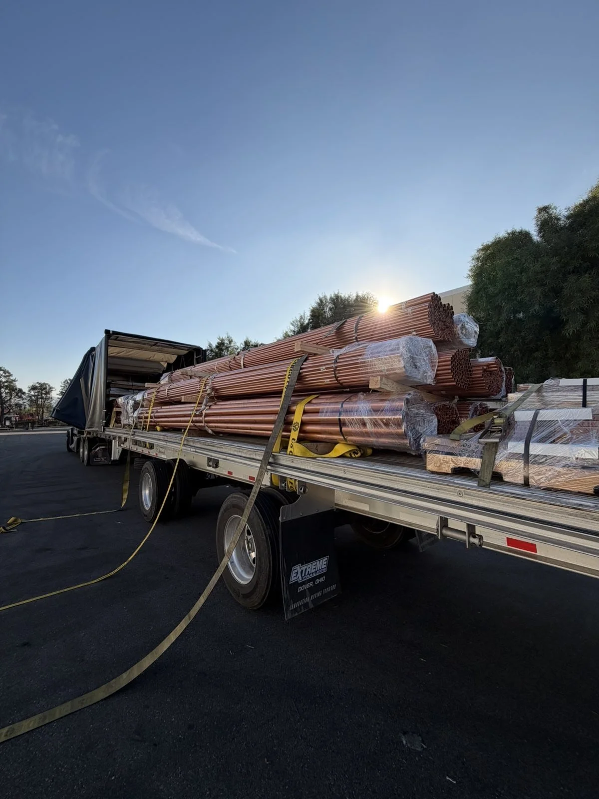 A large Link-Belt crane is being transported on a flatbed trailer along a highway under a blue sky with some wispy clouds.
