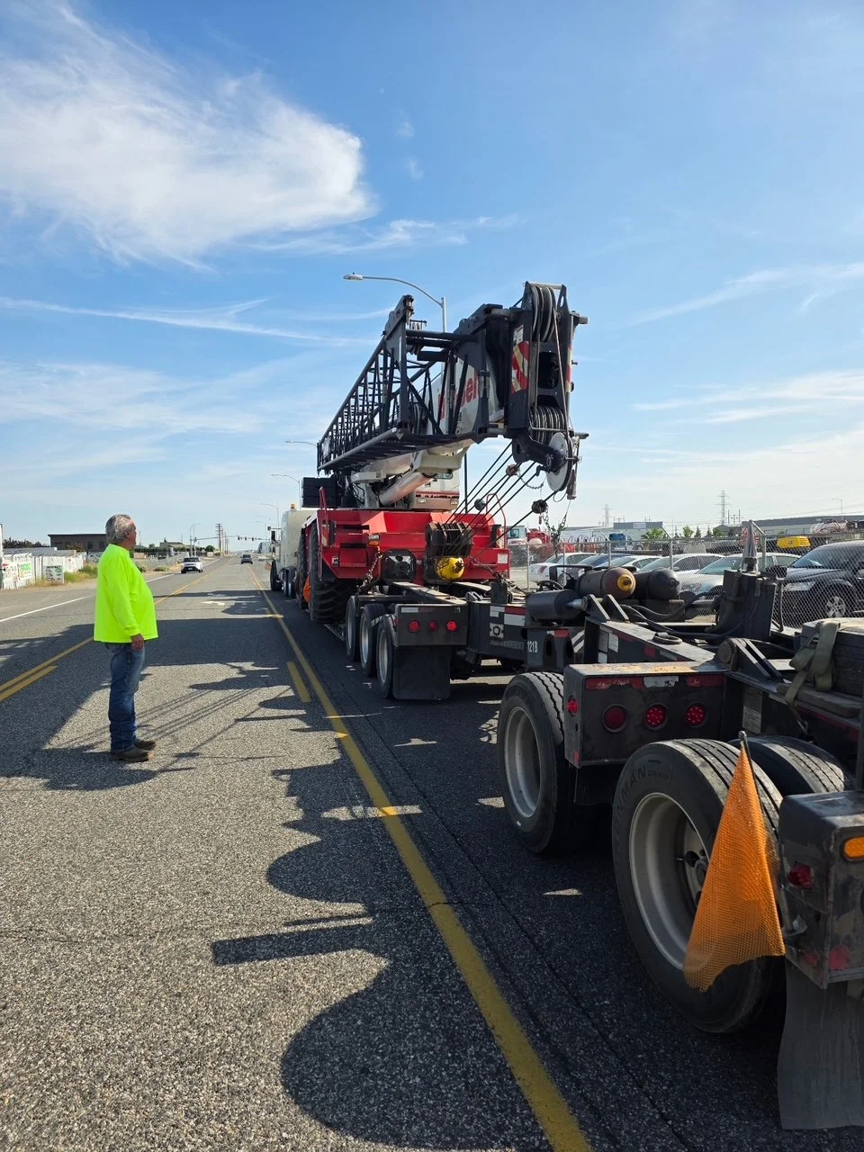 A large Link-Belt crane is being transported on a flatbed trailer along a highway under a blue sky with some wispy clouds.