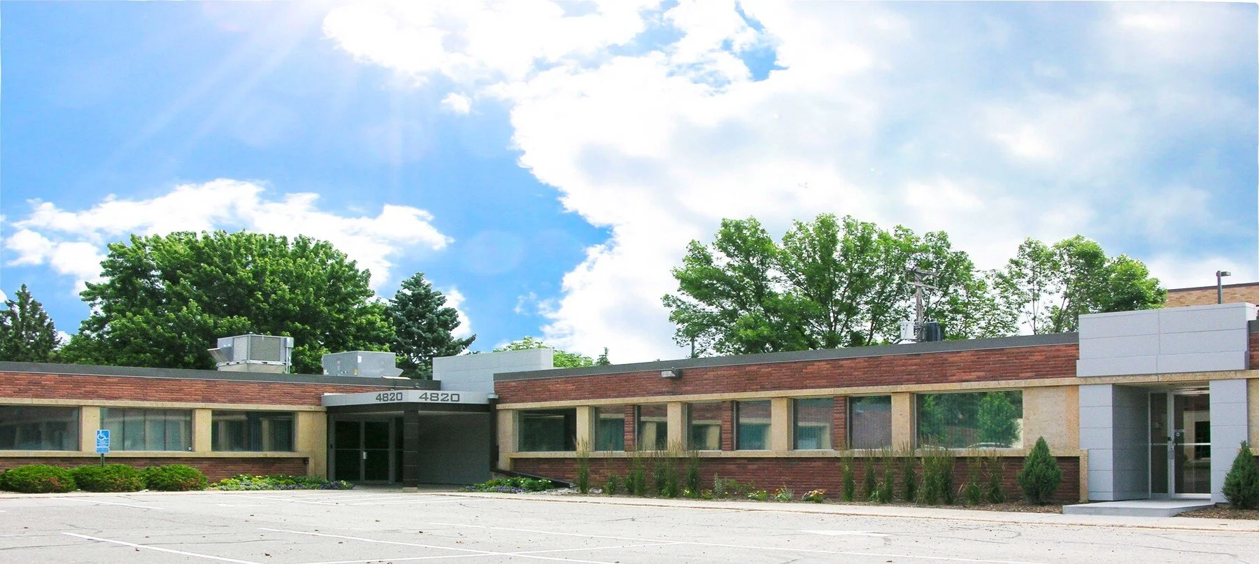 A pleasant, single-story office building with brick and beige exterior, large windows, and a parking lot in front. Trees and blue sky with clouds in the background.