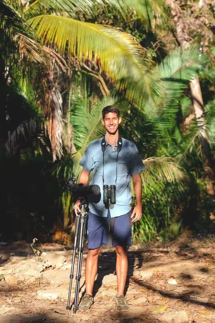 A group of hikers walking on a narrow trail through dense green forest. The person in the front is smiling and carrying a tripod on his shoulder, wearing a light-colored shirt and dark shorts.