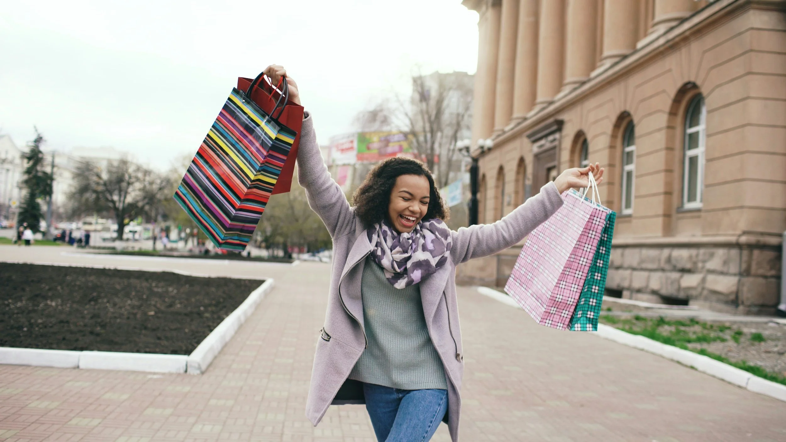 A young Black lady holding two shopping bags in each hand smiling and walking downtown.
