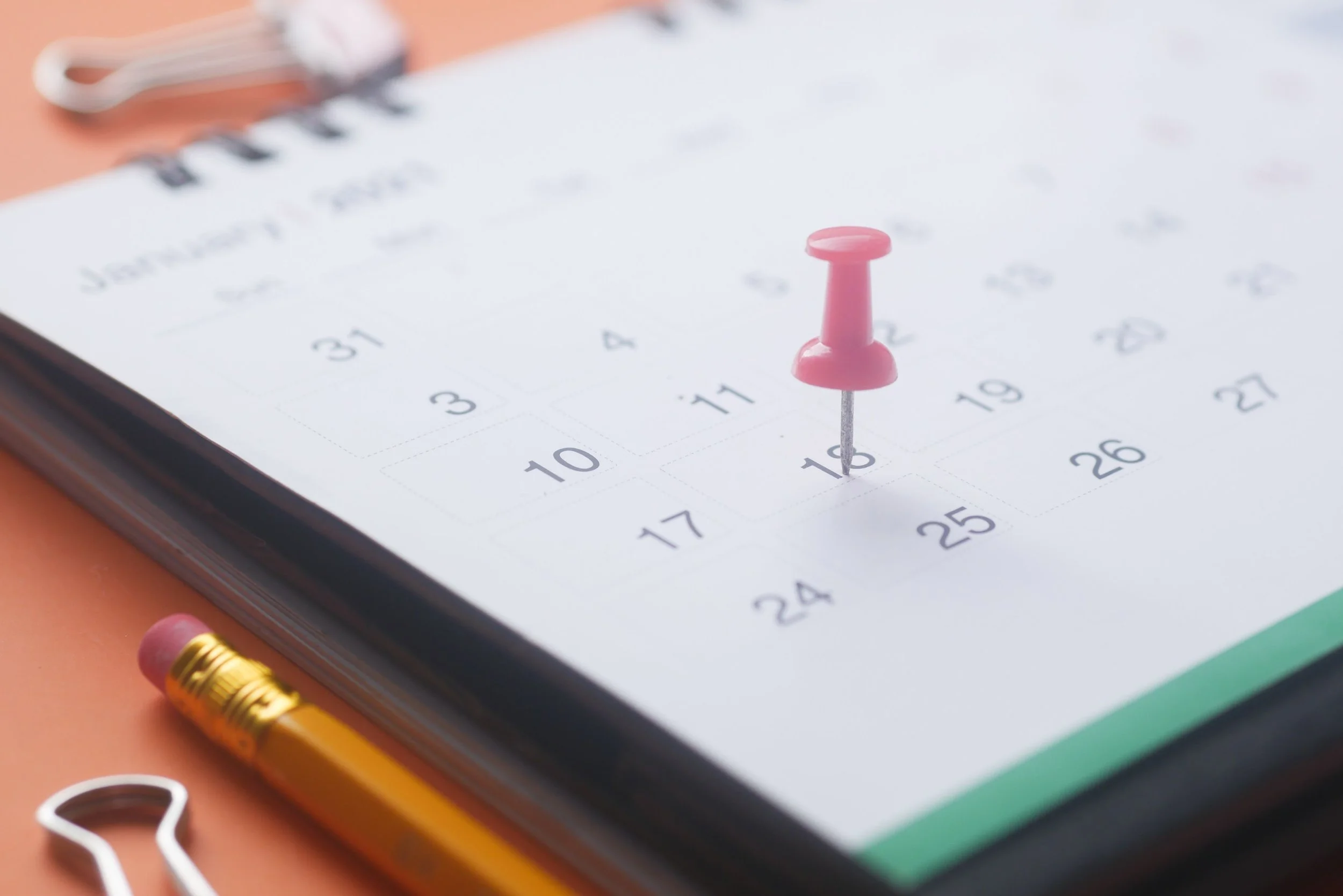 A pink push pin on a calendar displaying the date of November 15th. Nearby, there is a yellow pencil with an eraser, some paper clips, and a pair of scissors.