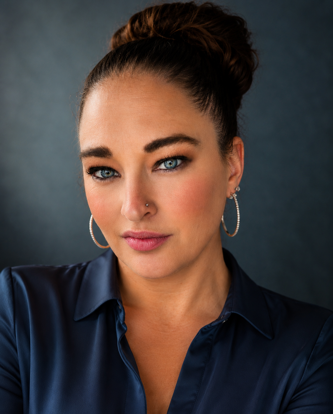 A woman with blue eyes, dark eyebrows, and a nose piercing, wearing large hoop earrings and a dark blue satin shirt, looking directly at the camera with a confident expression.